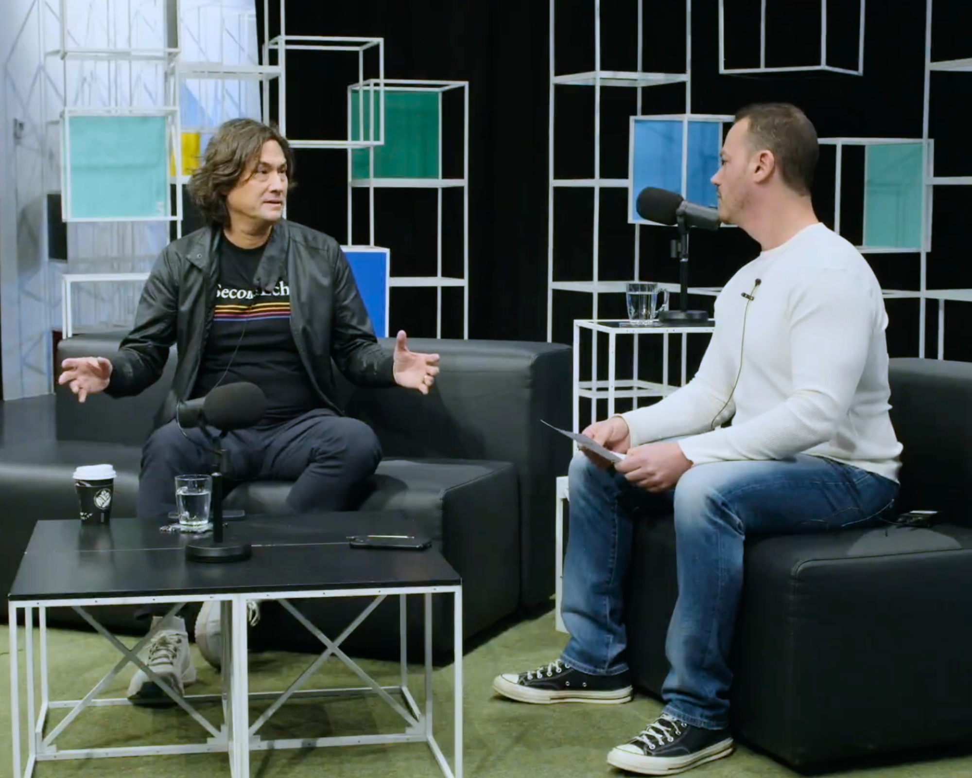 Two men engaged in conversation in a modern indoor setting with decorative cube-shaped shelves in the background. One man is sitting on a black couch, gesturing with his hand, and wearing a black leather jacket and a black T-shirt. The other man is standing, holding a paper, and wearing a white long-sleeve shirt and jeans. There are cups on the table in front of them, and a microphone is near the man sitting. The scene appears to be a recording or interview setup.