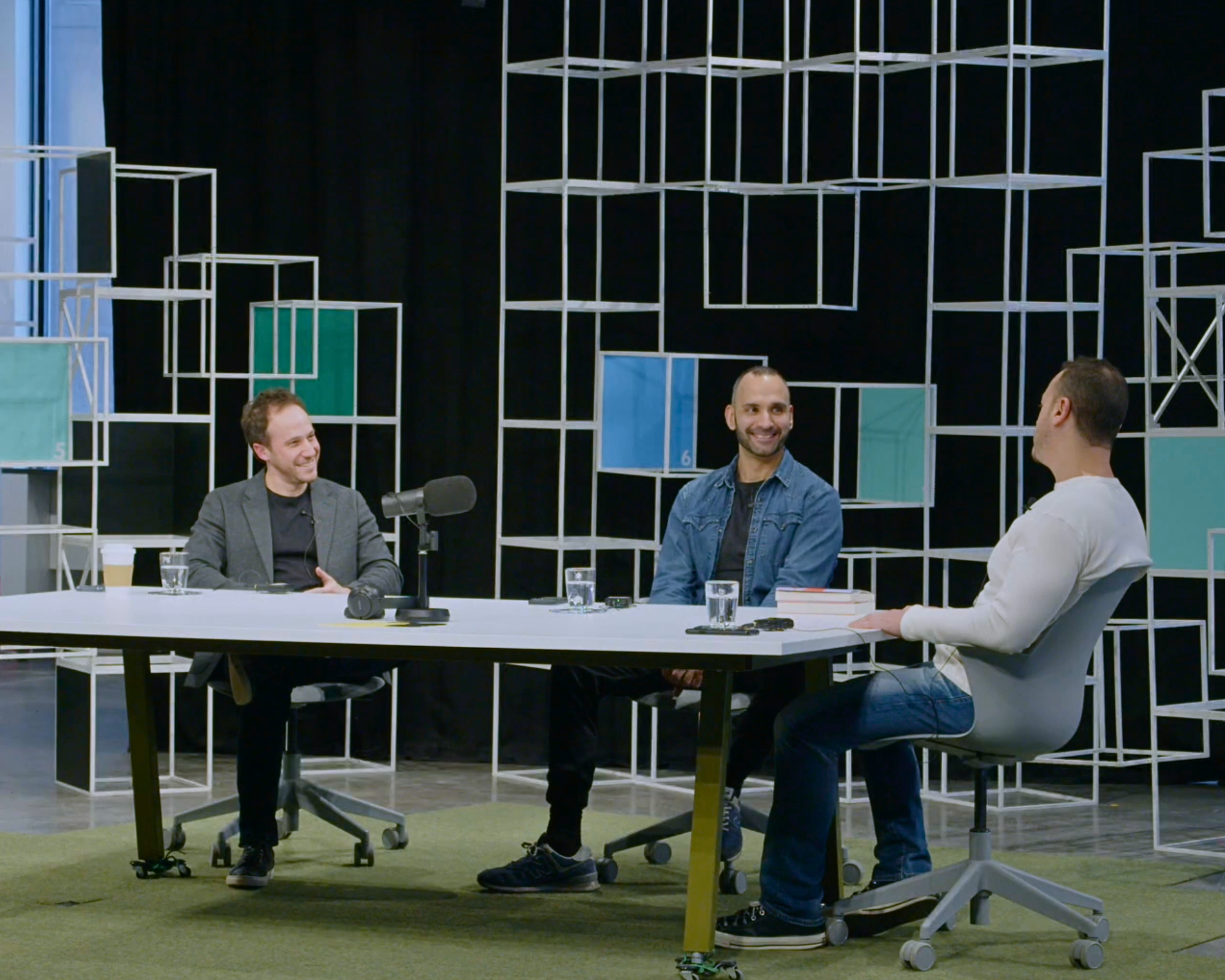 People sitting at a round table in a modern office or conference room, smiling and conversing, with a black backdrop and geometric wall design in the background.