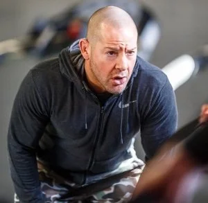 A man with a bald head and light skin, wearing a dark zip-up hoodie, looking surprised or confused.
