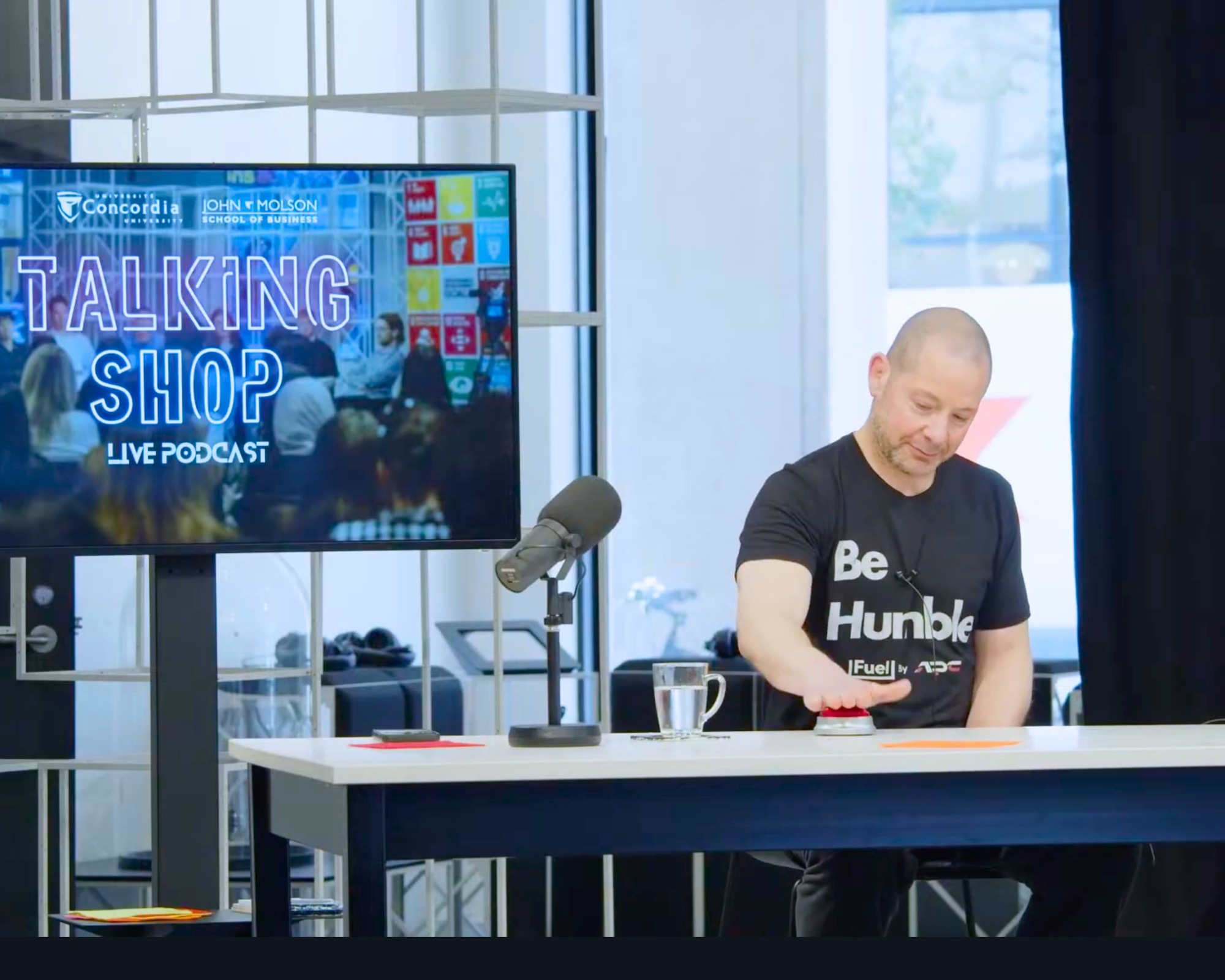 A man with a shaved head wearing a black T-shirt with white text, standing at a table, operating a red electronic device with a screen, in a modern room with a large flat-screen monitor displaying 'Talking Shop' on a TV stand to his left.