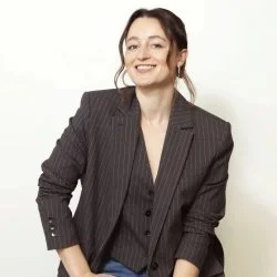 A woman smiling, wearing a black pinstripe blazer and dark shirt, standing against a white background.