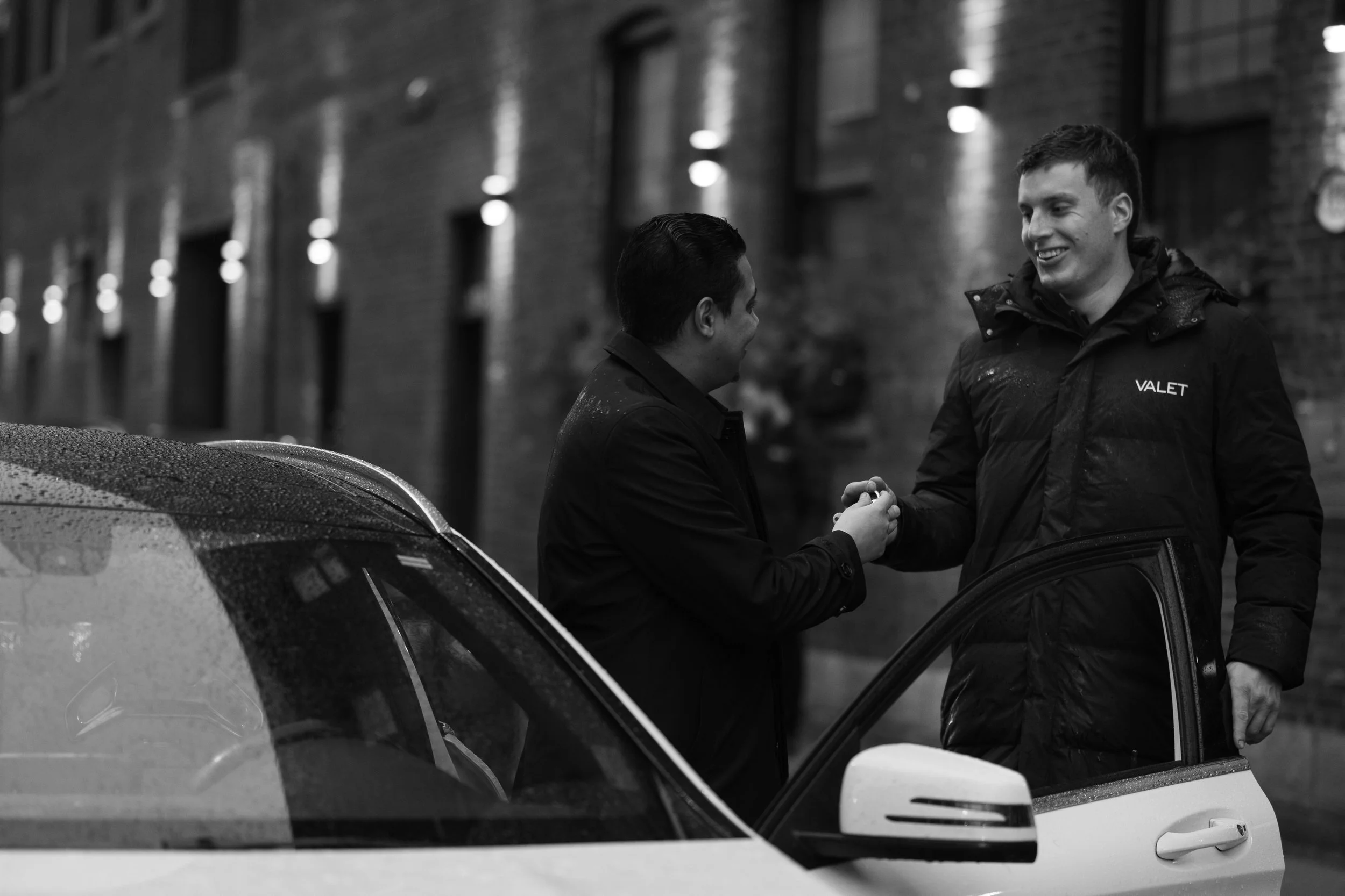 Two male valets shake hands on a rainy city street at night, with brick buildings and illuminated windows in the background.