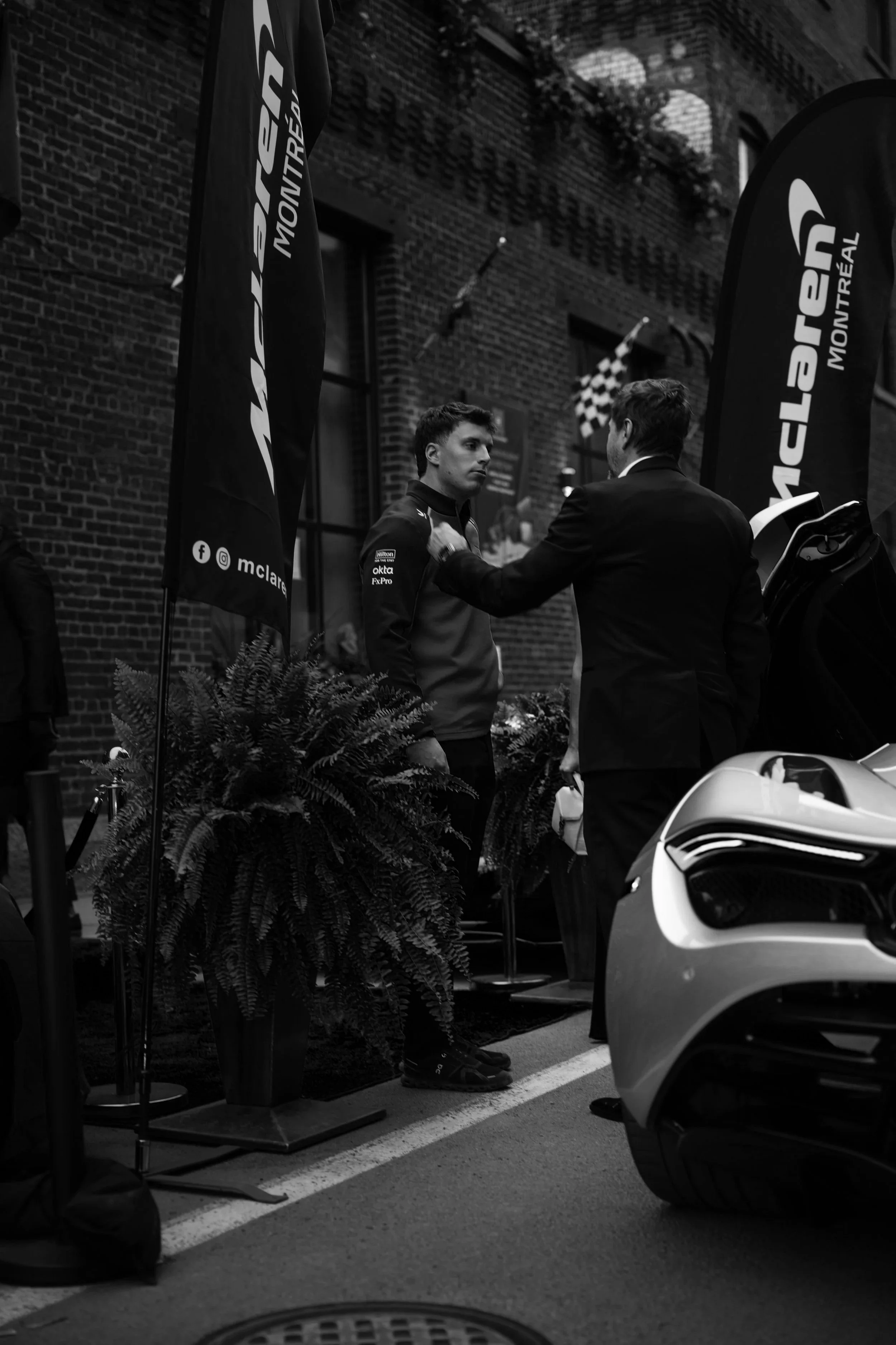 A man in a racing suit and a man in a suit are having a conversation outdoors, with flags, plants, a brick building, and a sports car visible in the background.