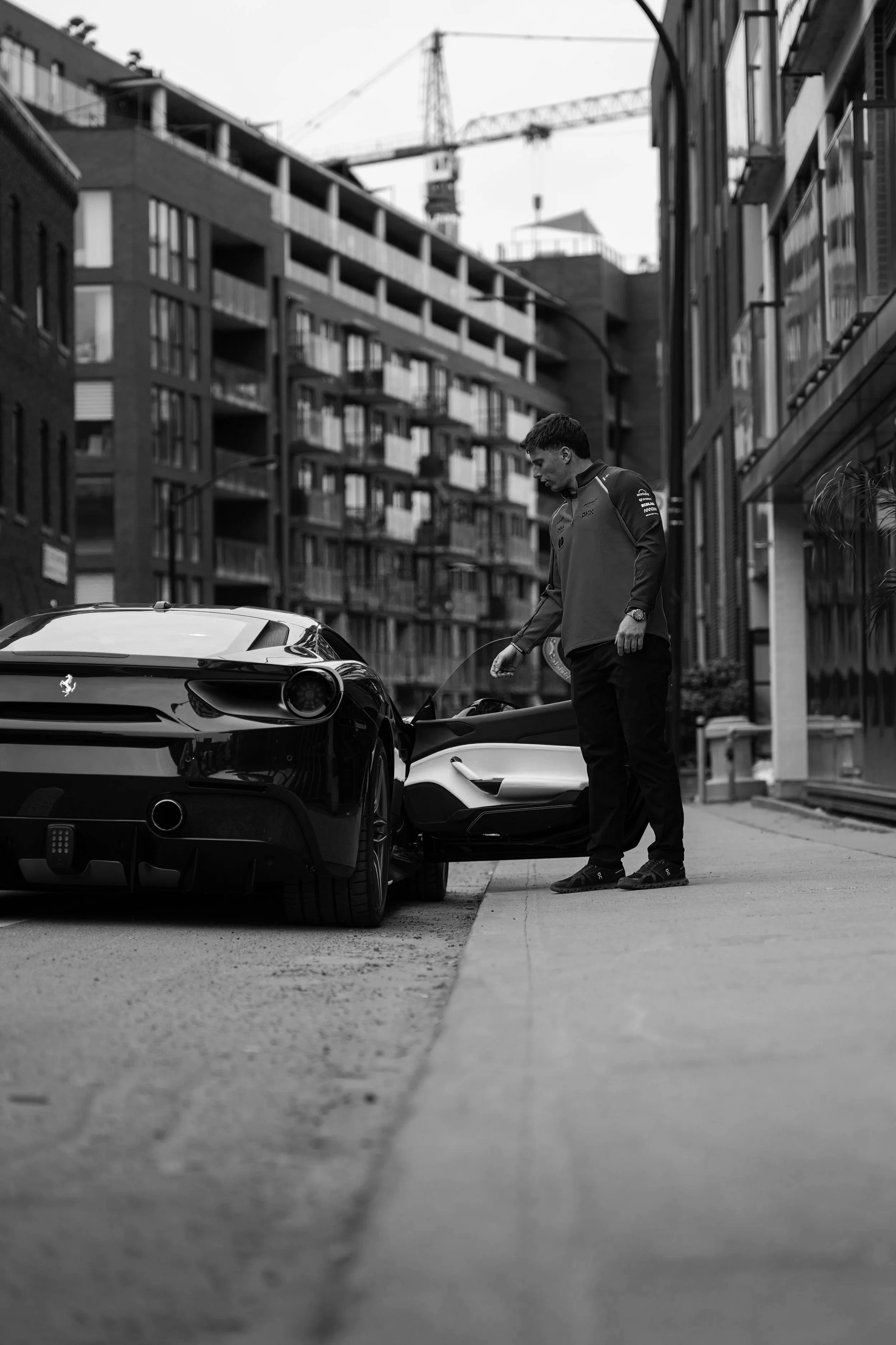 A man in a racing jacket standing next to two sports cars on a city street, with modern multi-story buildings and a construction crane in the background.