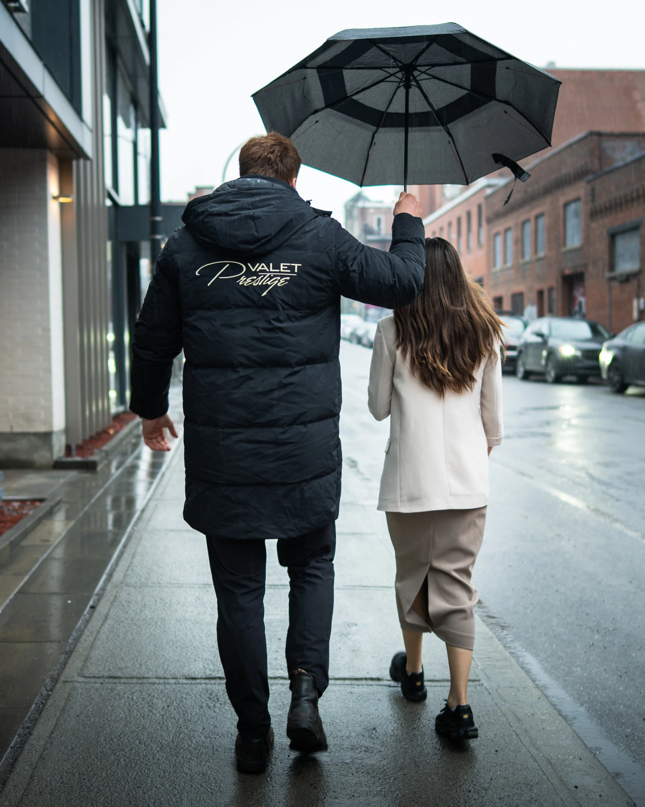 A man and woman walk together on a city sidewalk in rainy weather. The man holds a black umbrella and wears a dark jacket with 'VALET Prestige' on the back, while the woman wears a light-colored coat and black shoes.