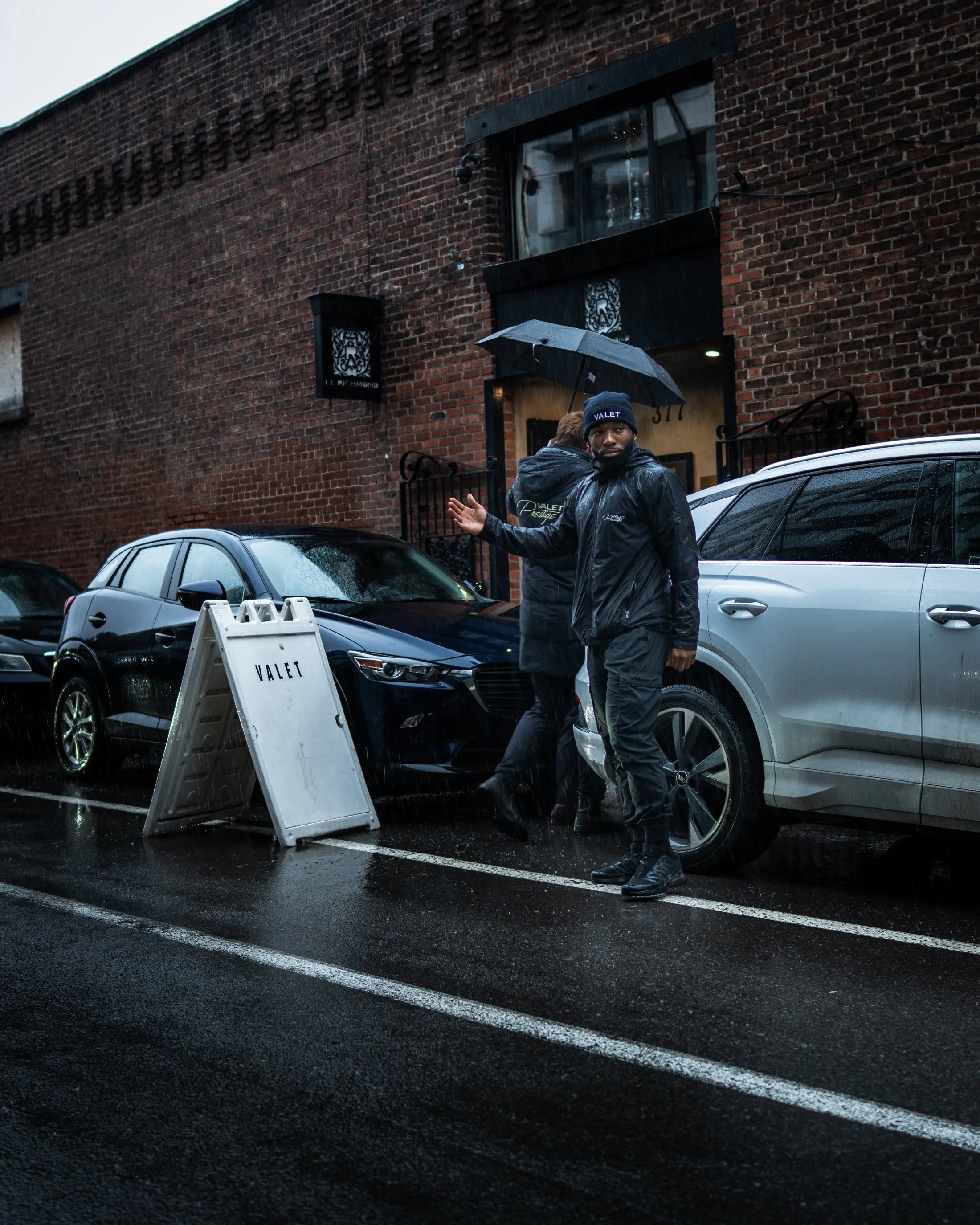 A man in a black jacket and black beanie with 'VALET' written on it stands in a parking lot holding a black umbrella, with cars parked nearby and a red brick building in the background on a rainy day.