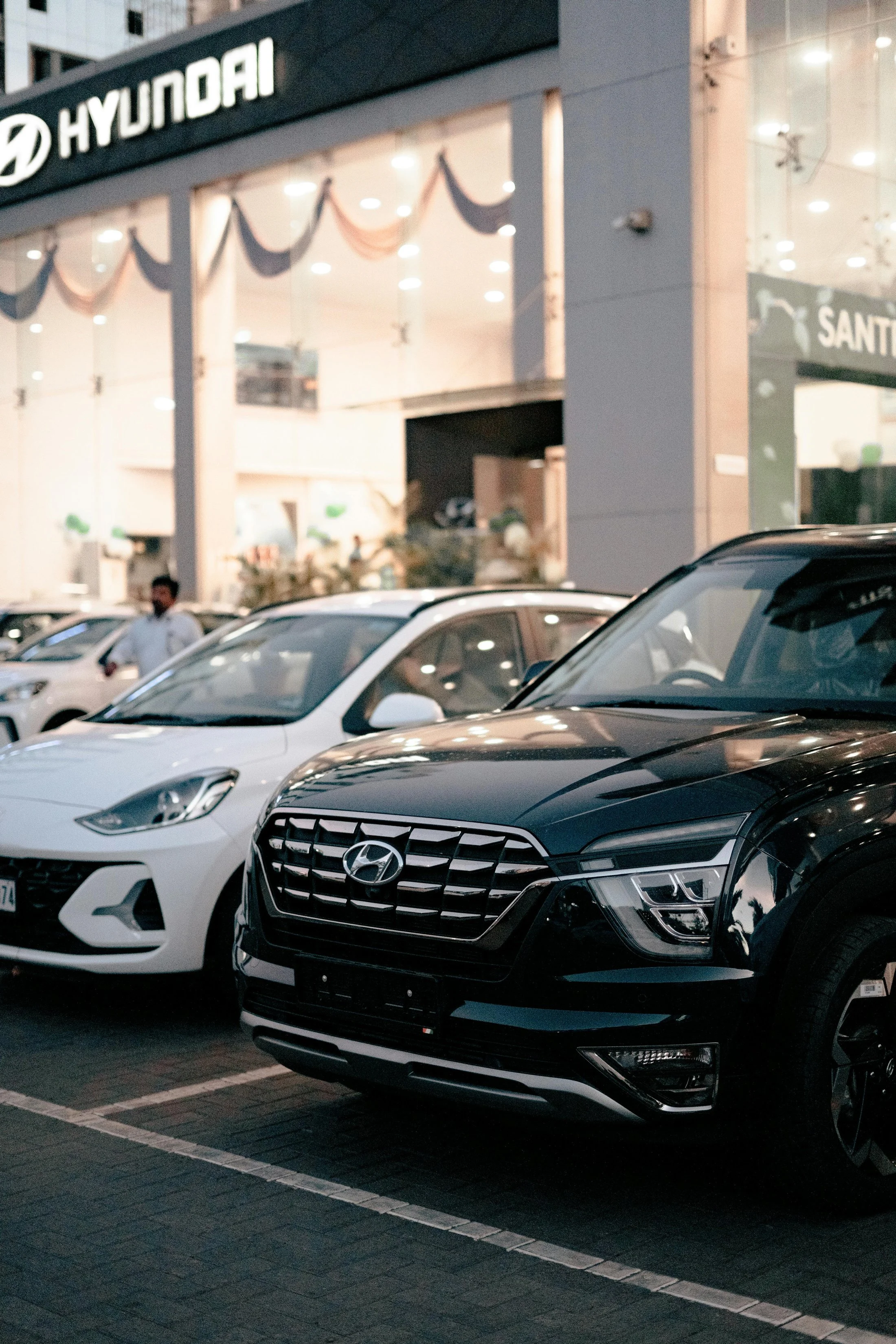 Parking lot with black and white Hyundai cars in front of a Hyundai dealership building decorated with drapes. There are lights on in the building and a person standing near the cars.