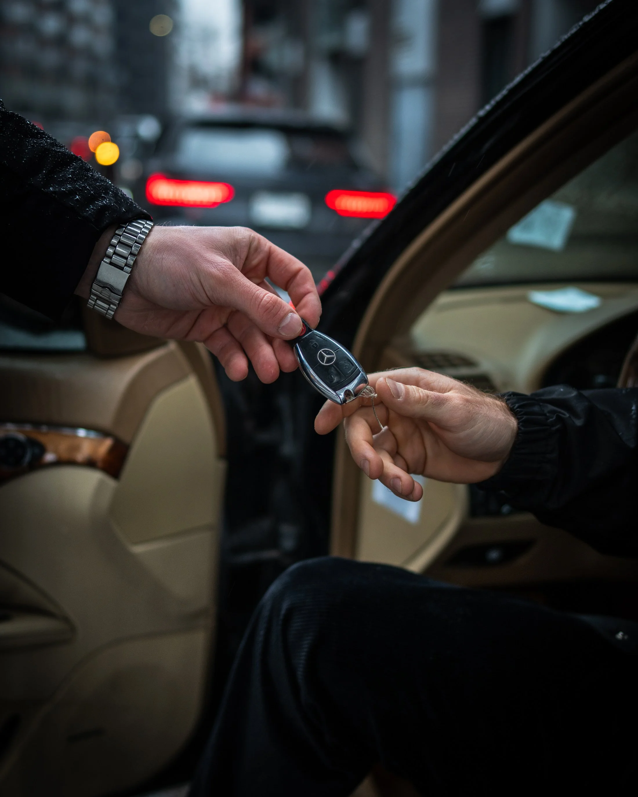 A person sitting in a vehicle on the passenger side, handing over a car key to another person. The key has a Mercedes-Benz logo. The scene is observed from the back seat of the vehicle, with a blurred view of other cars outside in traffic.