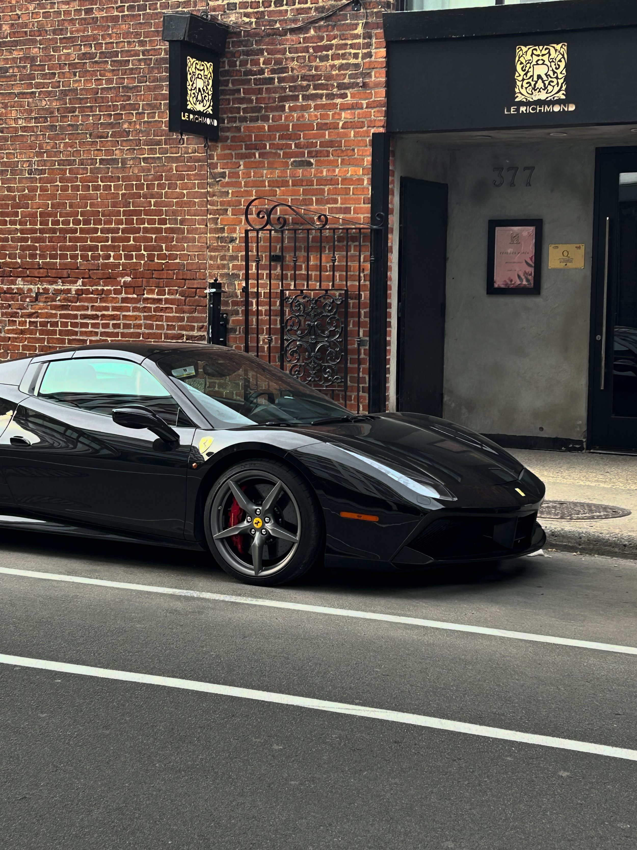 A black Ferrari sports car with red brake calipers parked on a city street, with a brick building and signage in the background.