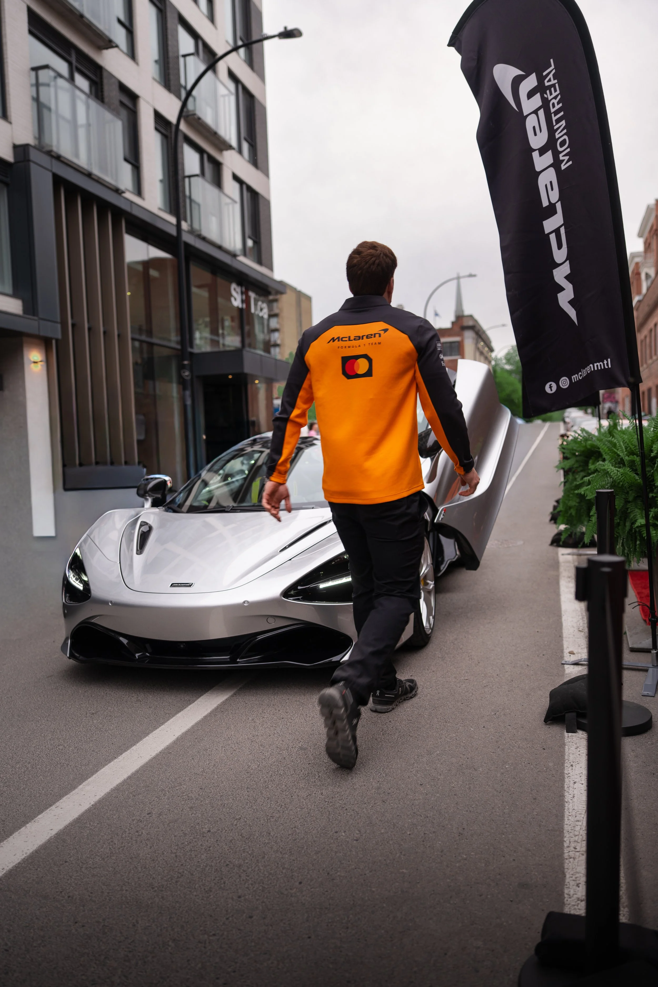 A man wearing a McLaren team jacket is walking towards a silver sports car with upward-opening doors on a city street. There is a black flag with white text that reads 'Wheel,' and the background shows modern buildings and street lamps on an overcast day.