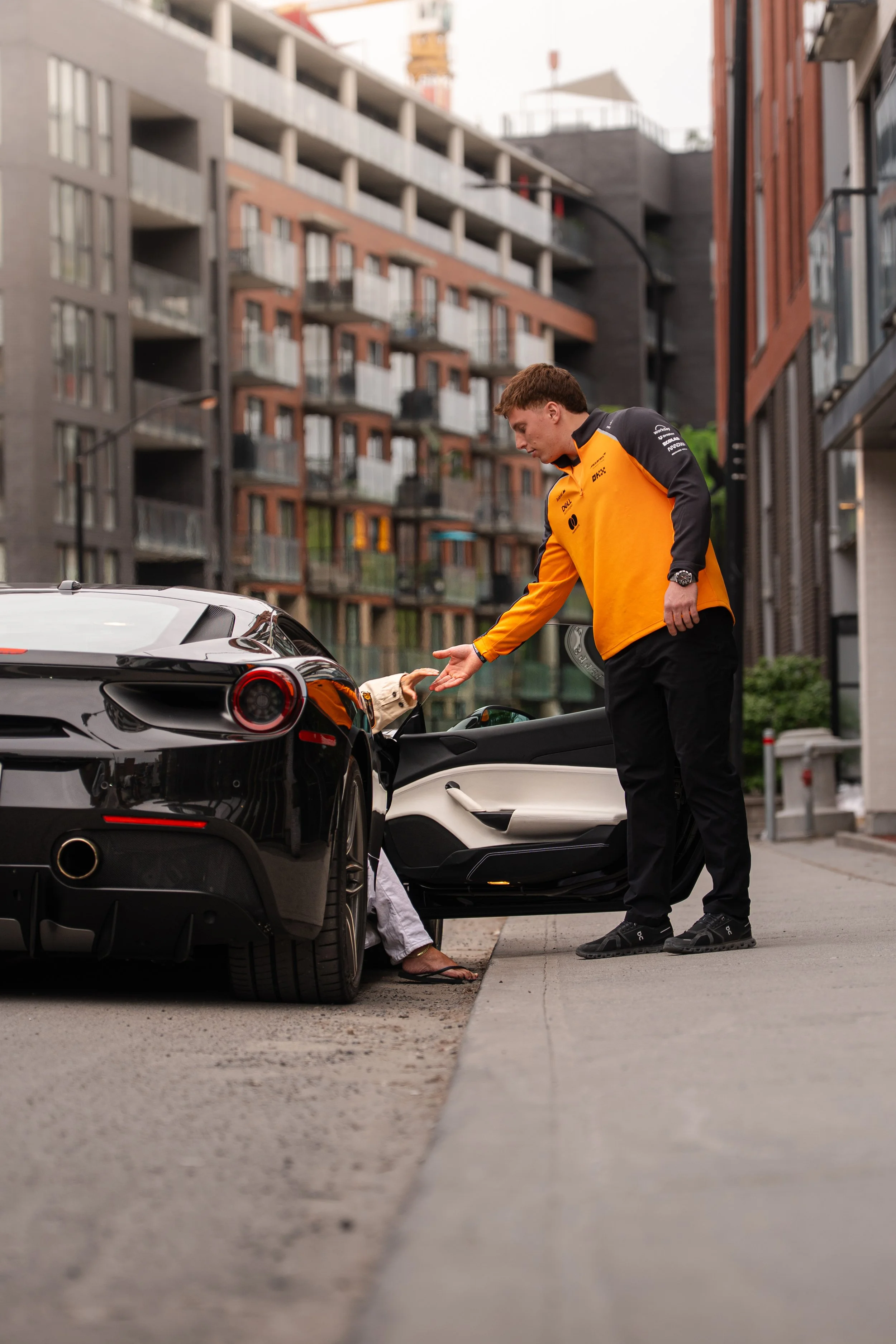 A man in a yellow racing jacket is seen exchanging money with a person inside a black sports car parked on the street, with modern apartment buildings in the background.