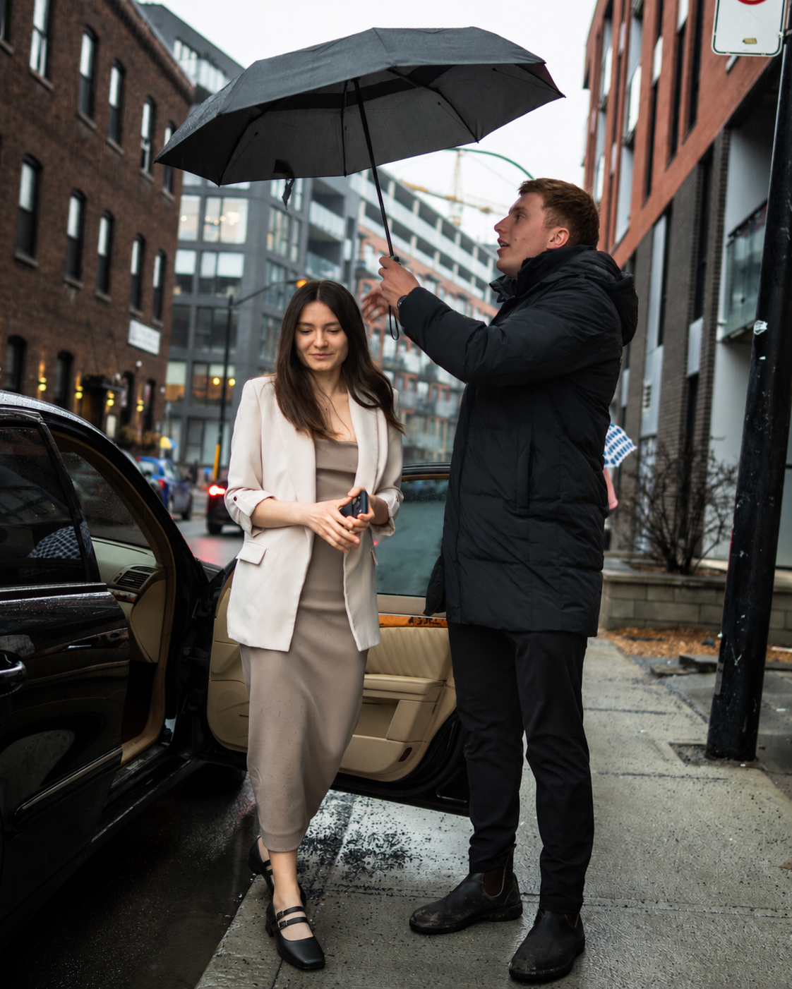 A man holding an umbrella over a woman as they stand beside a black car on a rainy street, with modern buildings in the background.