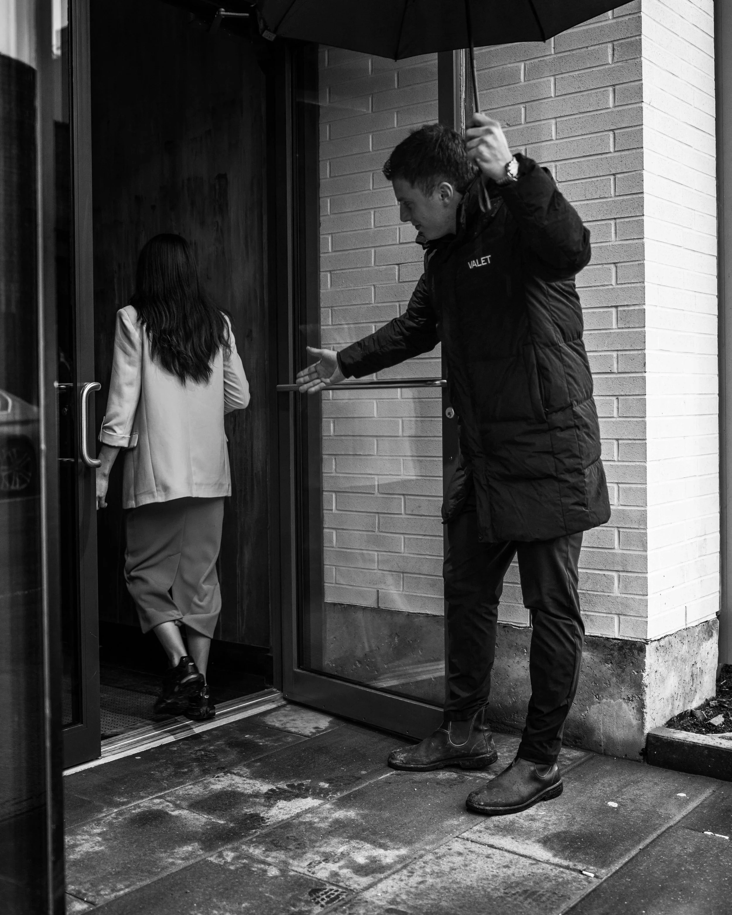 A valet in dark clothing holding an umbrella for a woman in a coat entering a building.