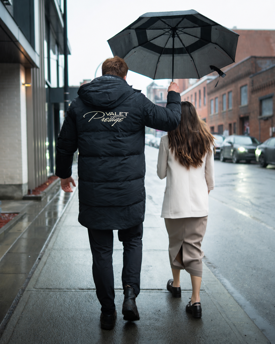 A man and woman walking on a rainy city sidewalk, with the man holding an umbrella over both of them. The man wears a black jacket with "Valet Prestige" written on the back, and the woman wears a light-colored coat and skirt.