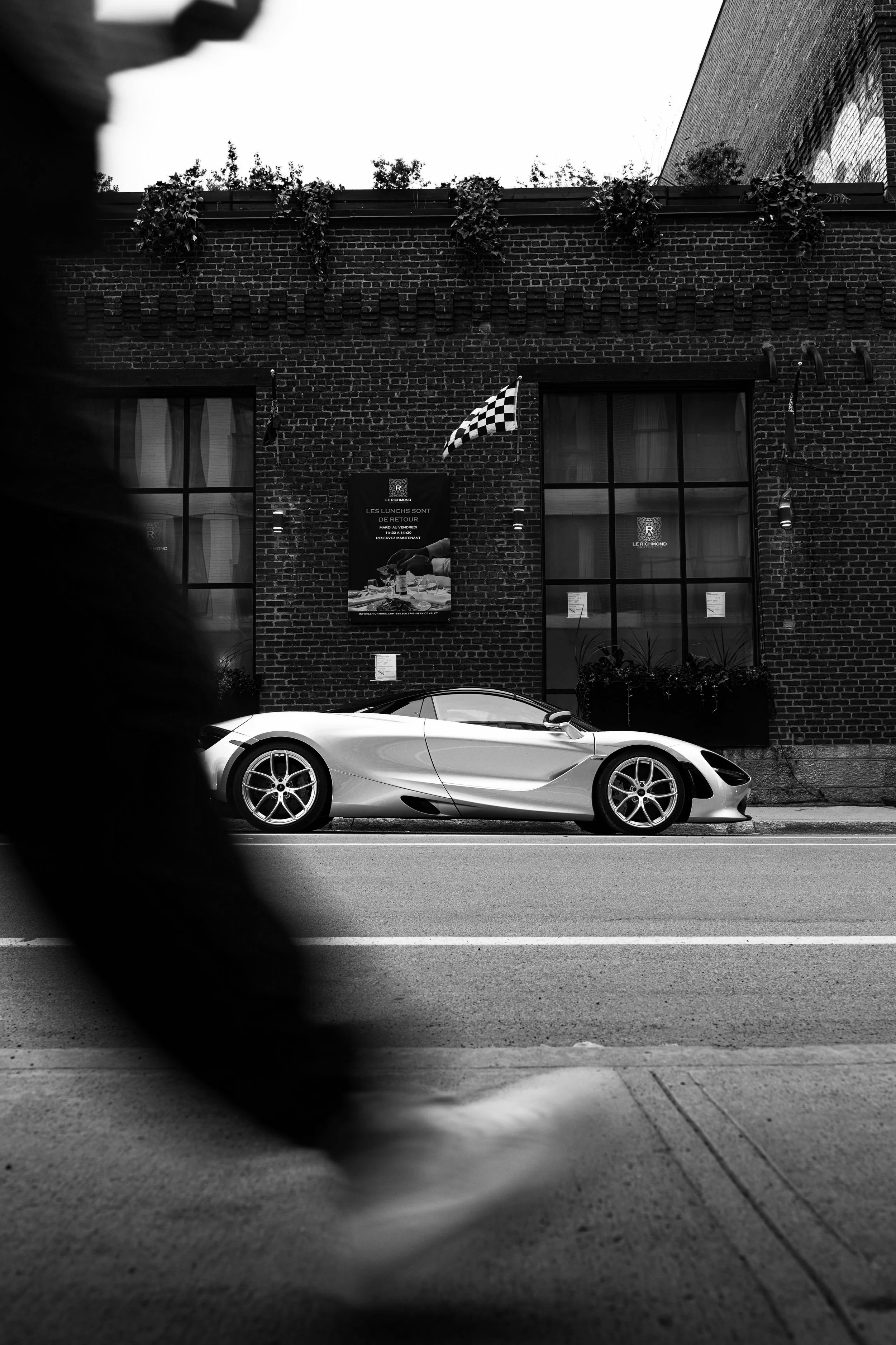A sleek silver sports car parked along a city street in front of a brick building with large windows. The photo is taken from a low angle, showcasing the car's modern design, with a blurred foreground and a brick building with chimneys and potted plants in the background.