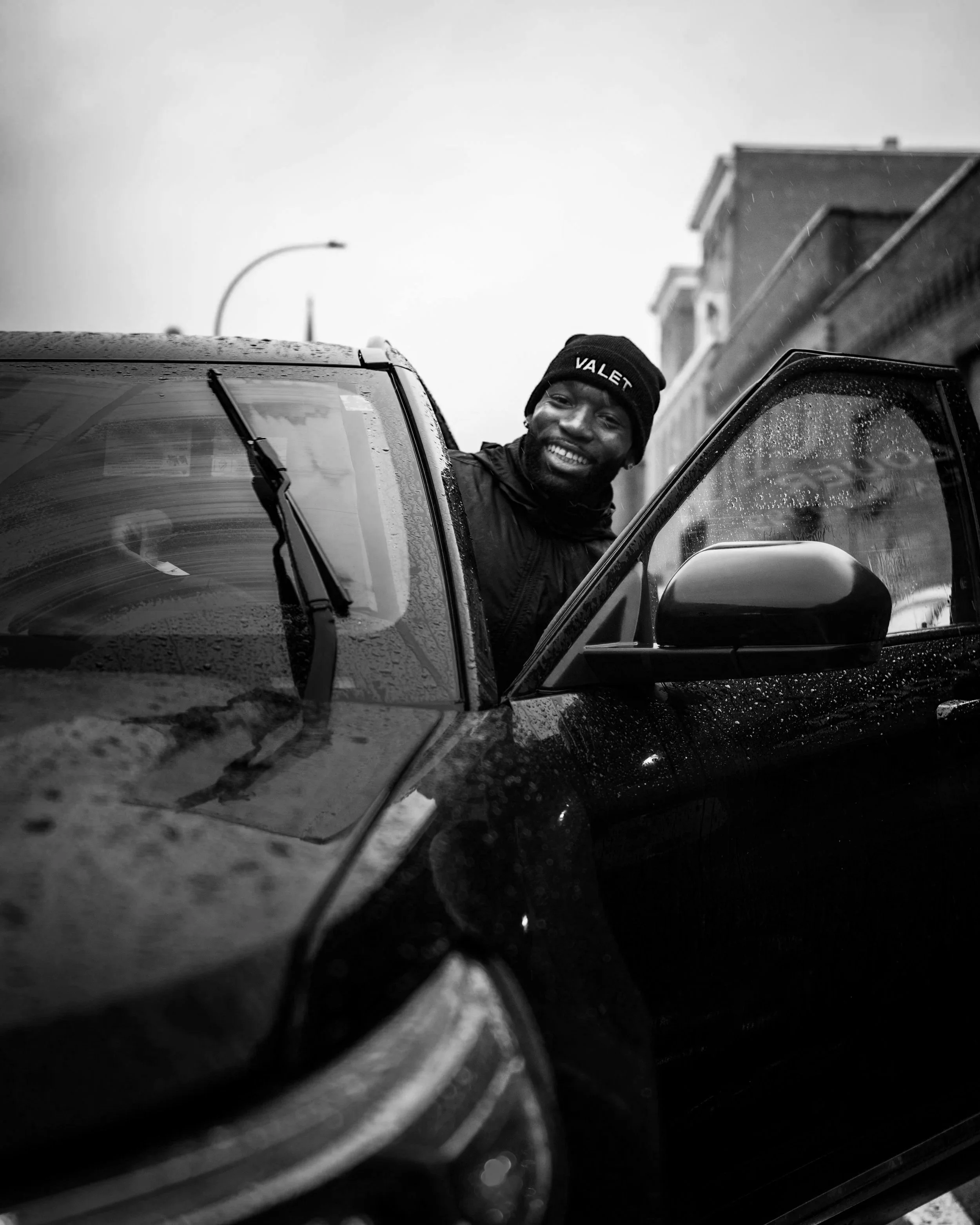 A smiling man wearing a beanie with 'VALET' embroidered on it, leaning out of the driver side window of a wet car, on a city street with residential buildings and a streetlamp in the background.
