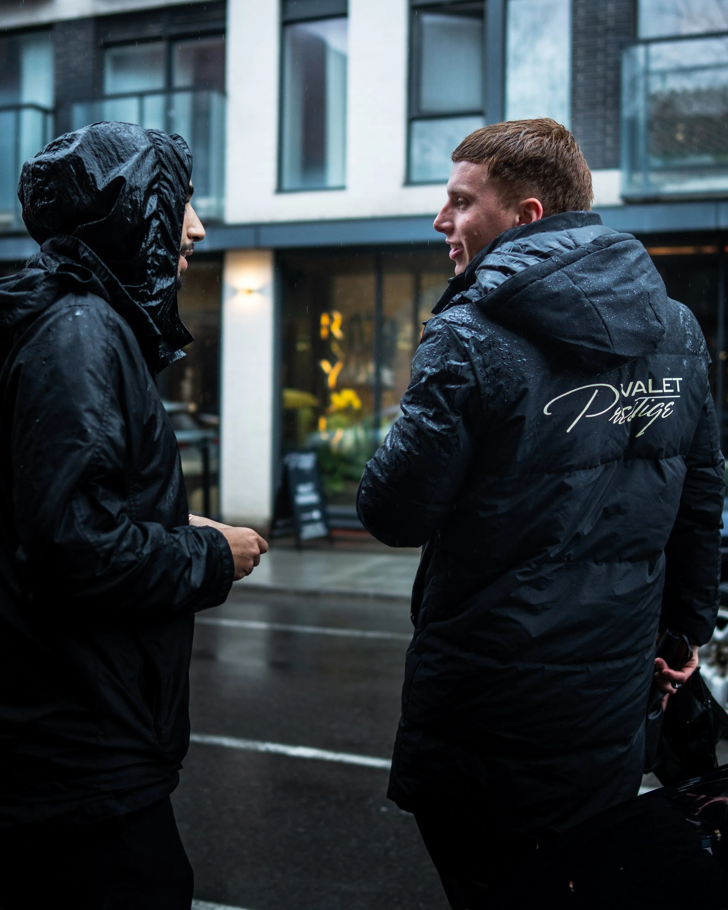 Two young men in black rain jackets facing each other and talking on a rainy street, with a modern building in the background.