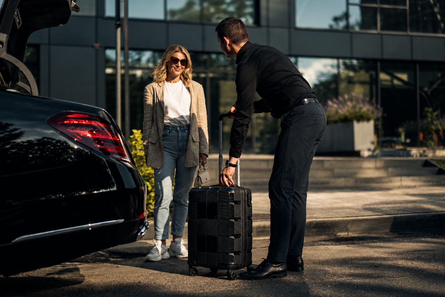 A man checking in luggage at a car with a woman standing nearby, outside a modern building, during daytime.