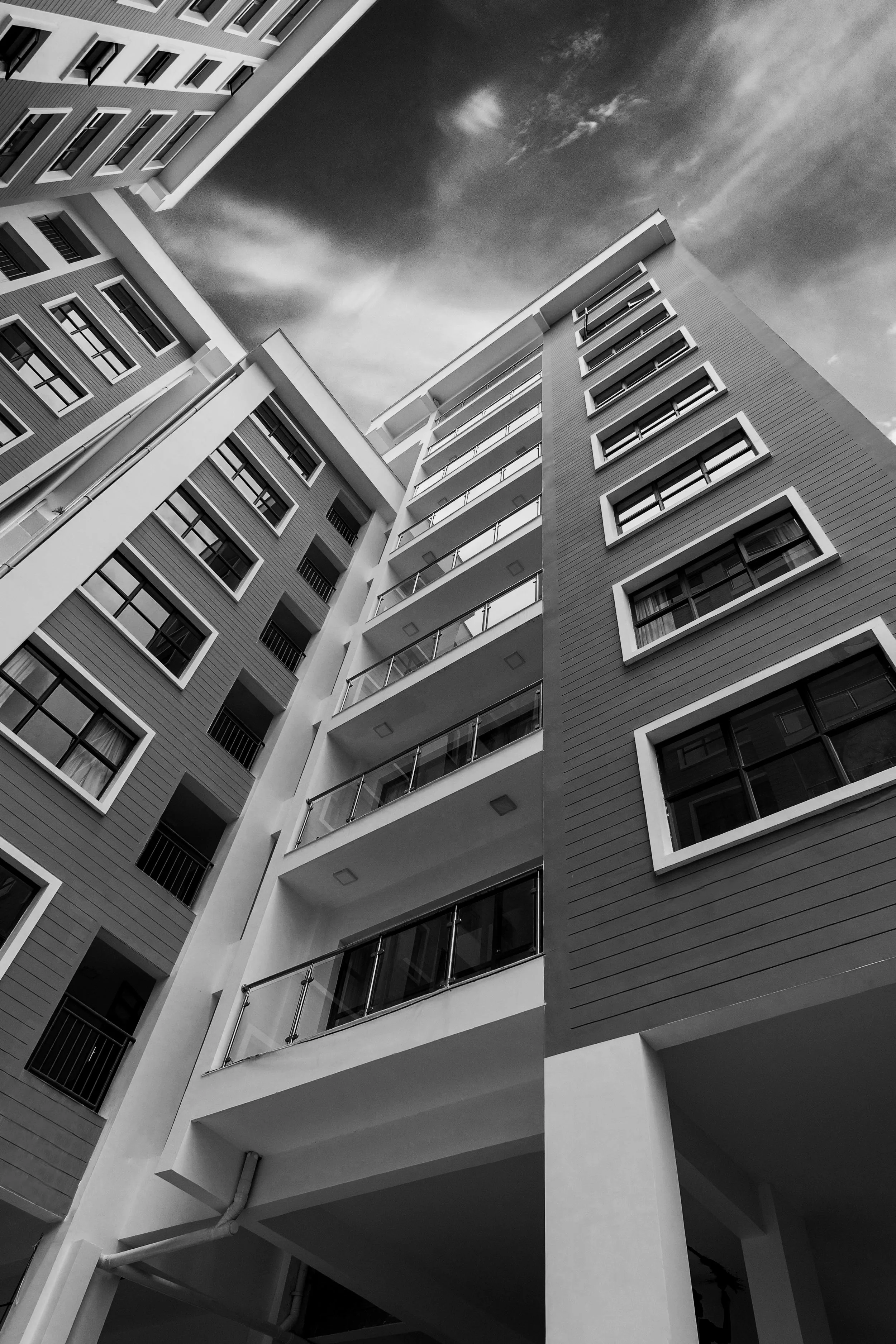 Black and white photo of modern multi-story apartment building with multiple balconies, numerous windows and a cloudy sky in the background.