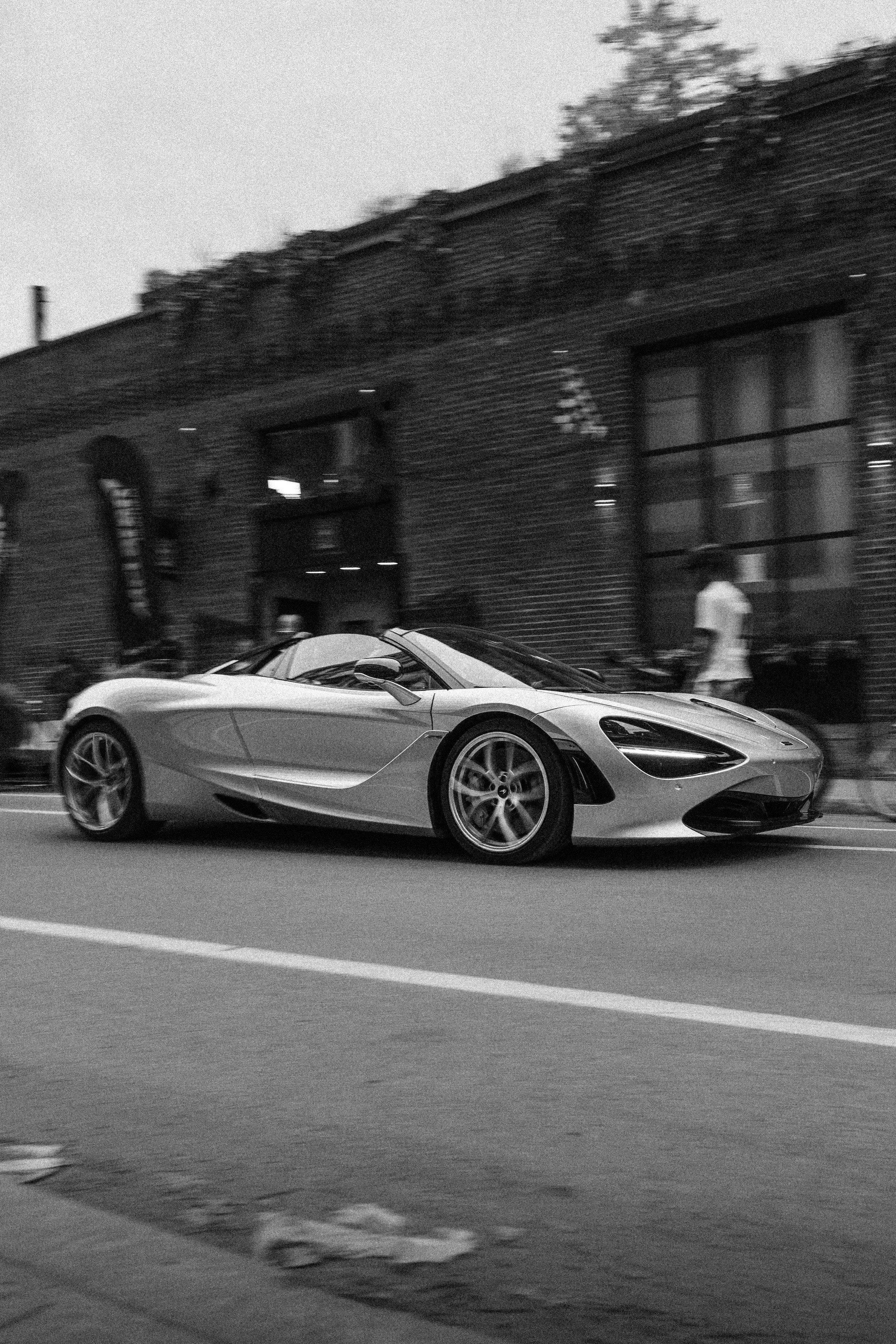 A white sports car is driving on a city street next to a brick building. A person is walking on the sidewalk nearby, and the cars are captured in motion.