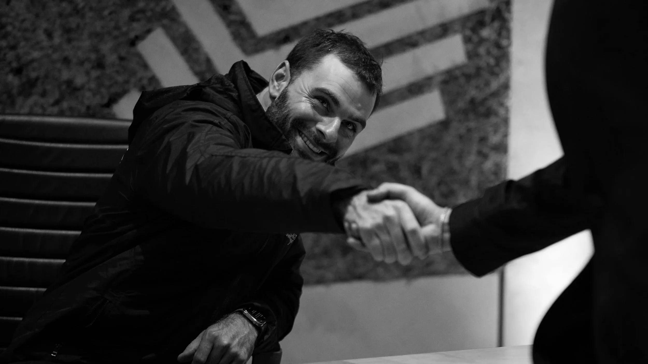 A man with a beard smiling and shaking hands with another person, sitting on a bench against a wall with stairs in the background.