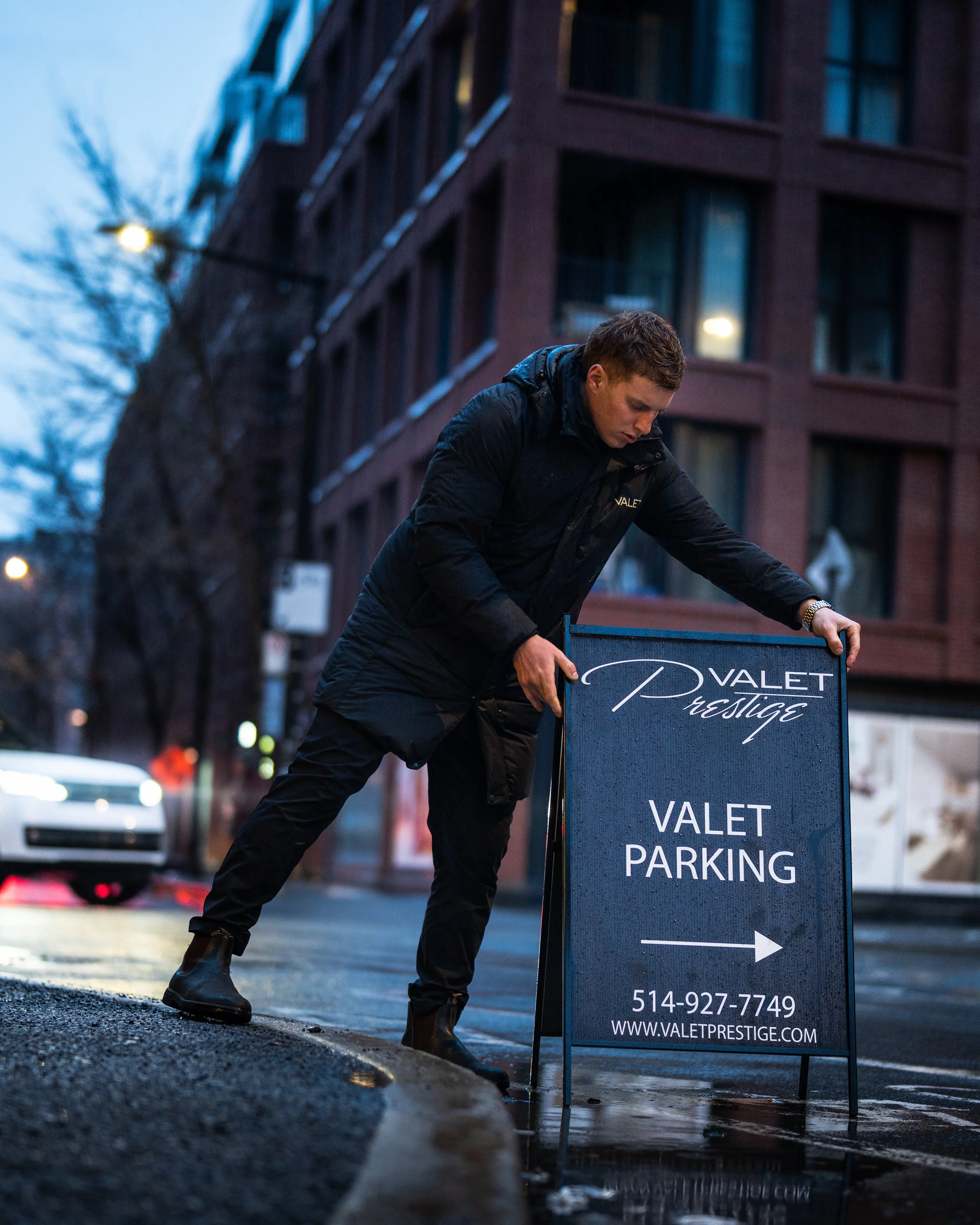 A man, wearing a black jacket and black boots, is adjusting a valet parking sign on a wet city street during evening or early night. There is a building with lit windows in the background, and the street is illuminated by streetlights and vehicle headlights.