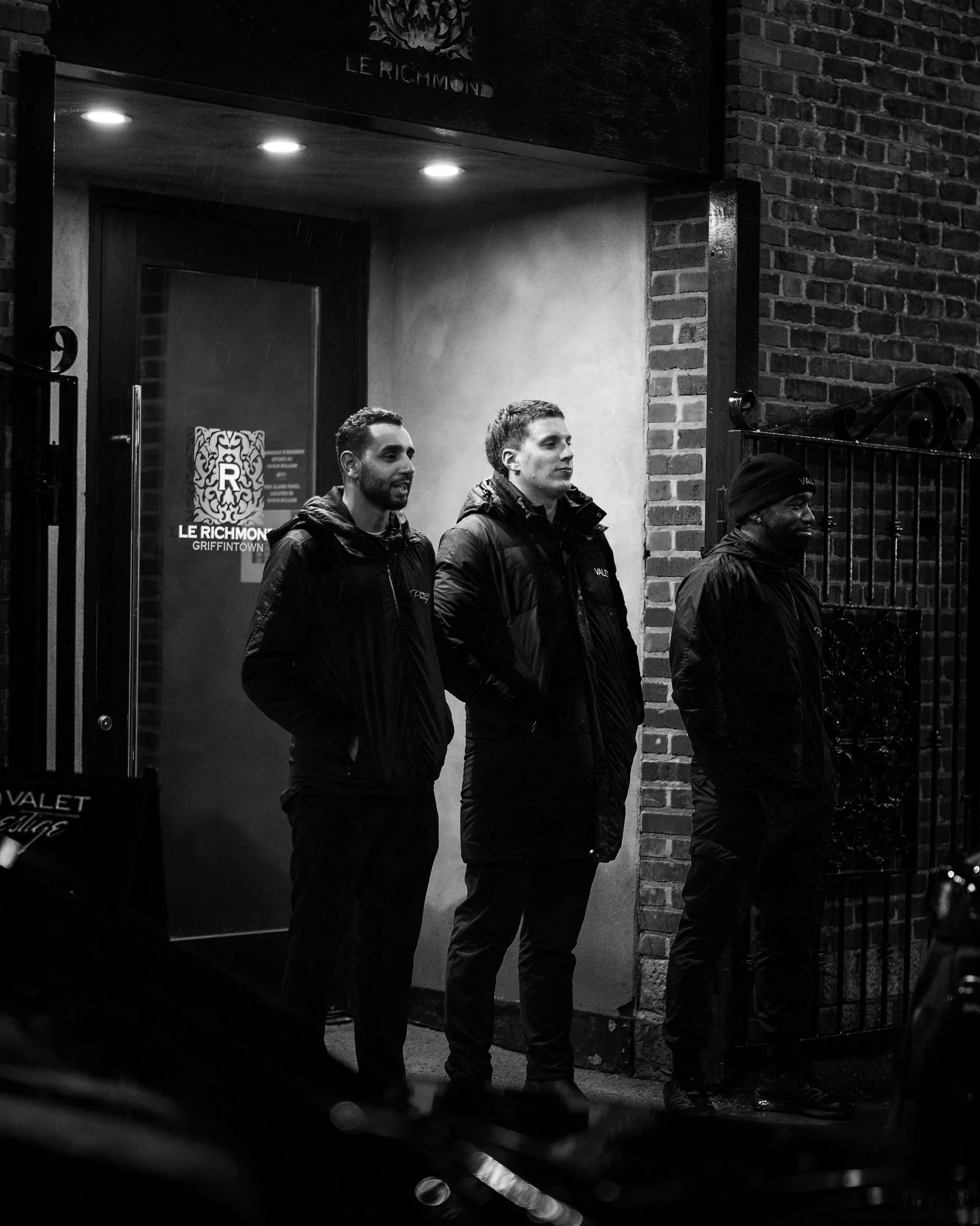 Three young men standing outside the entrance of Le Richmond Griffin Town at night, with a brick wall and gate nearby, illuminated by overhead lights, in black and white.