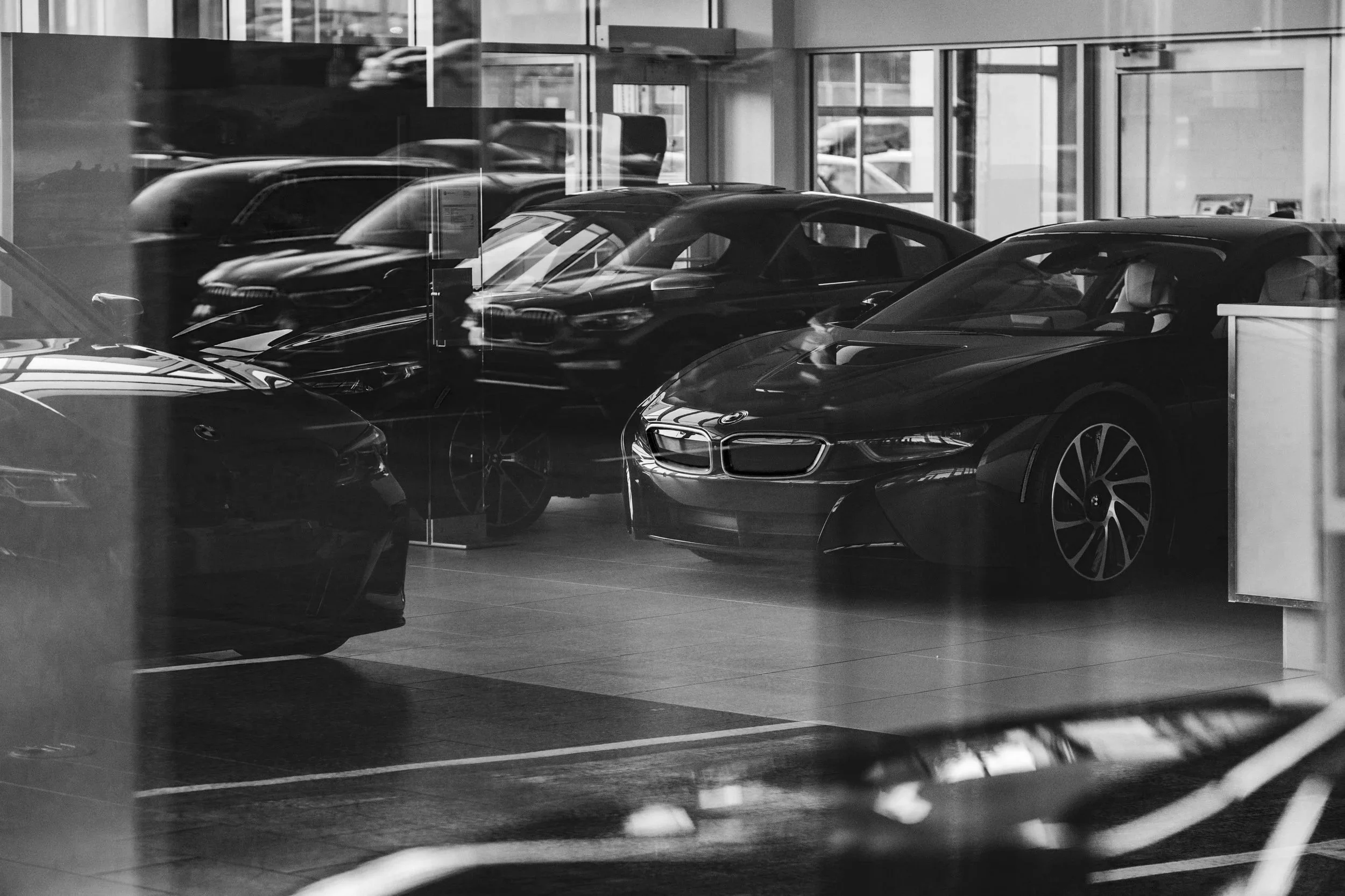 A black-and-white photo of a luxury car showroom with several sports cars on display behind glass. The cars have sleek designs and are arranged inside the showroom, visible through the glass window.