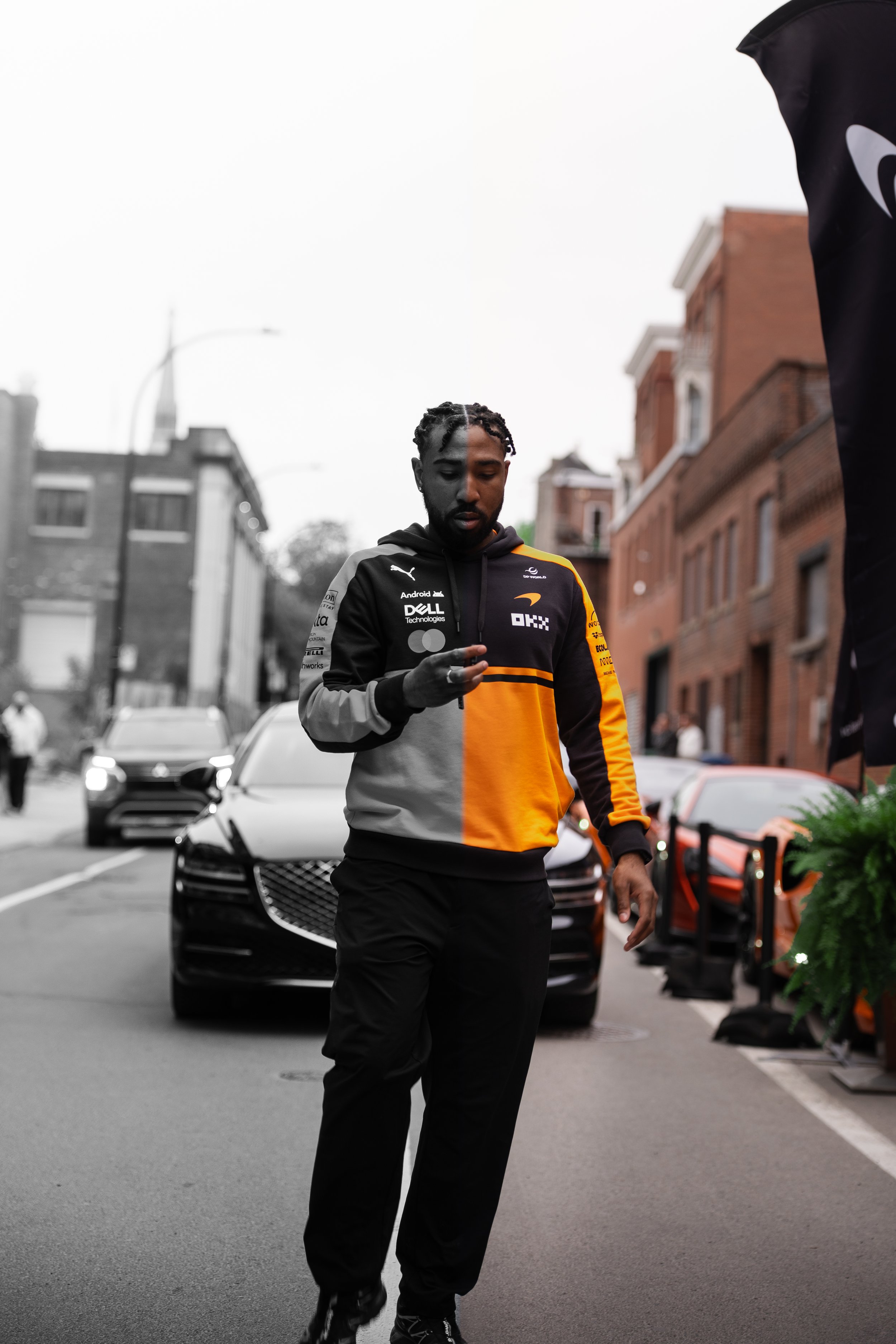 A man with a beard and braided hair, wearing a black and yellow sports jacket, is looking down at his phone while standing on a street. The background is mostly black and white, with cars and brick buildings visible. Part of a black flag or banner is on the right side of the image.