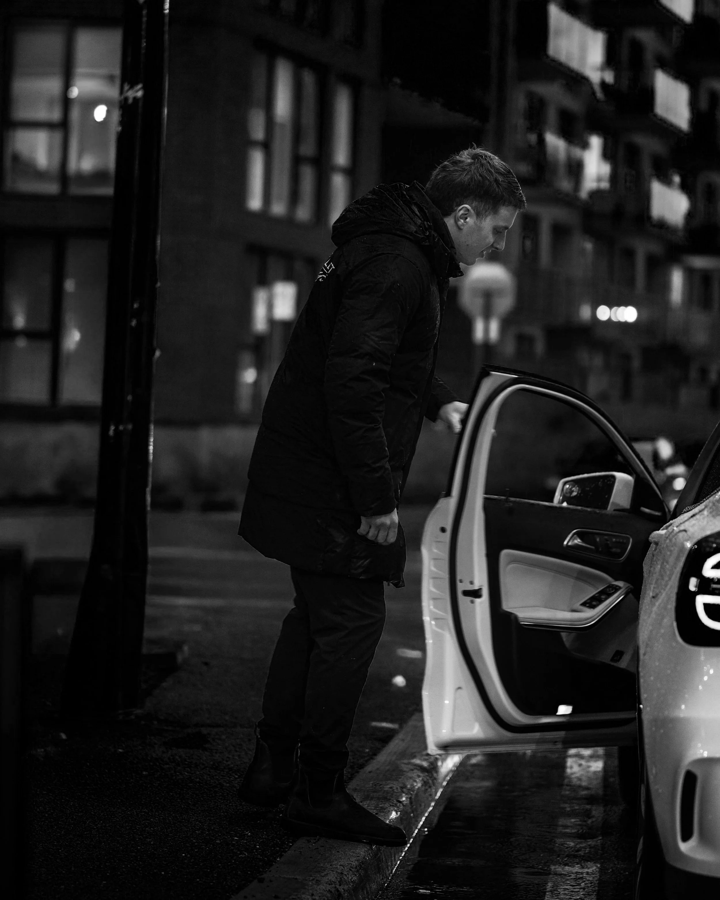A young man in a dark jacket stands next to an open car door during rainy night, looking down with a somber expression, in an urban setting with residential buildings in the background.