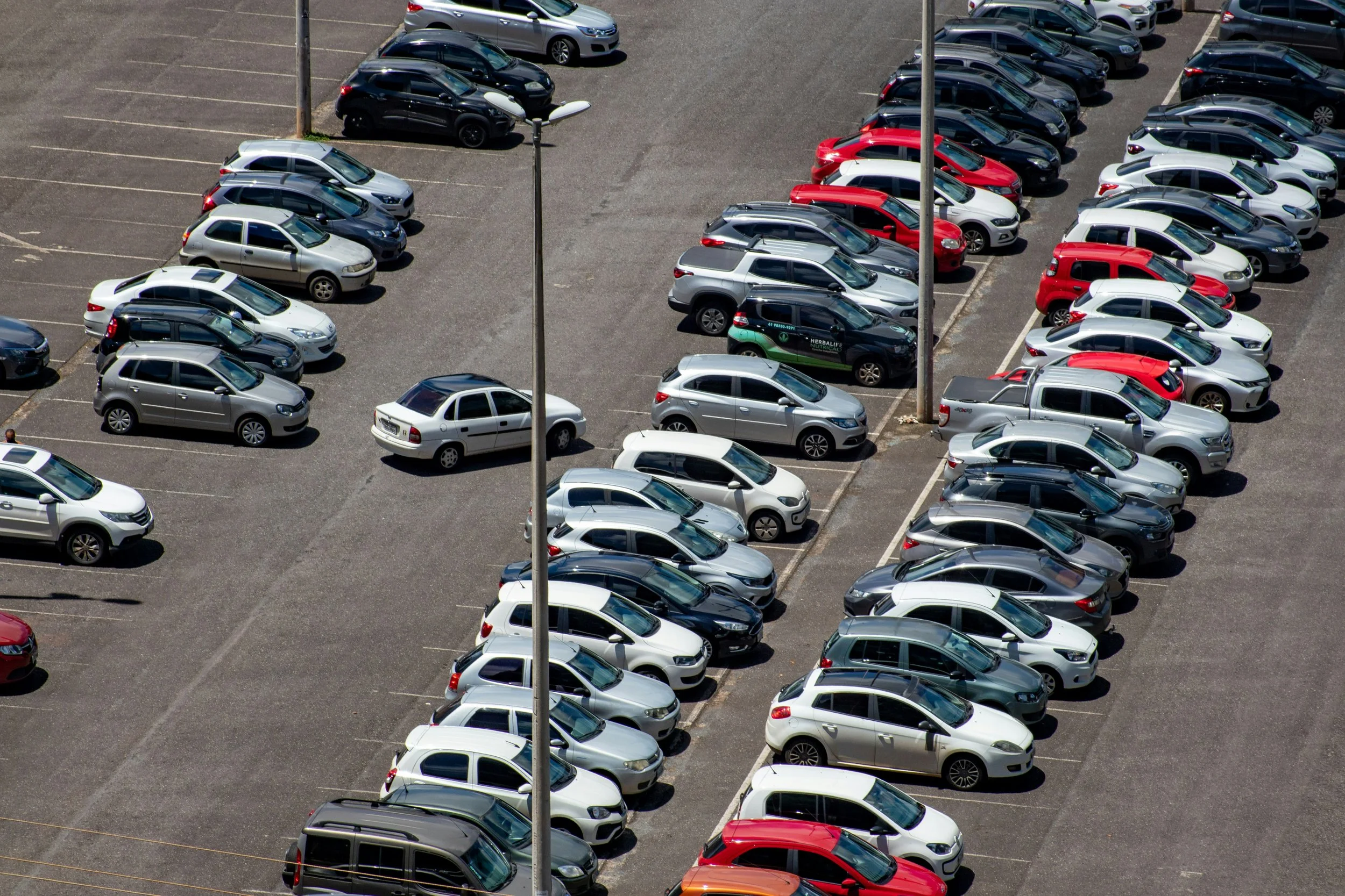 An aerial view of a parking lot filled with various cars including sedans, SUVs, and hatchbacks parked in designated spaces, with two streetlights visible in the image.