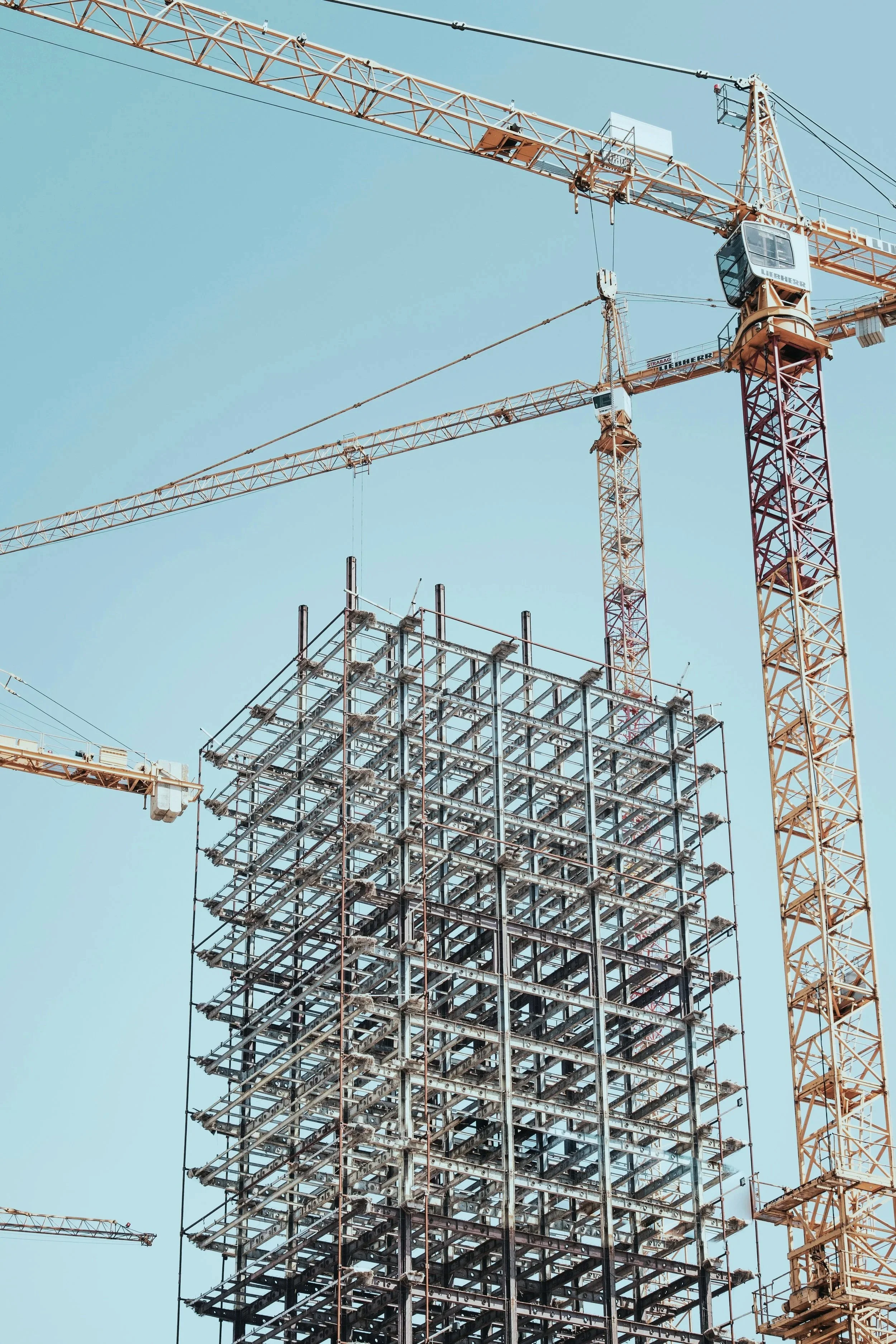 Two large construction cranes are working on a high-rise building under construction against a clear blue sky.