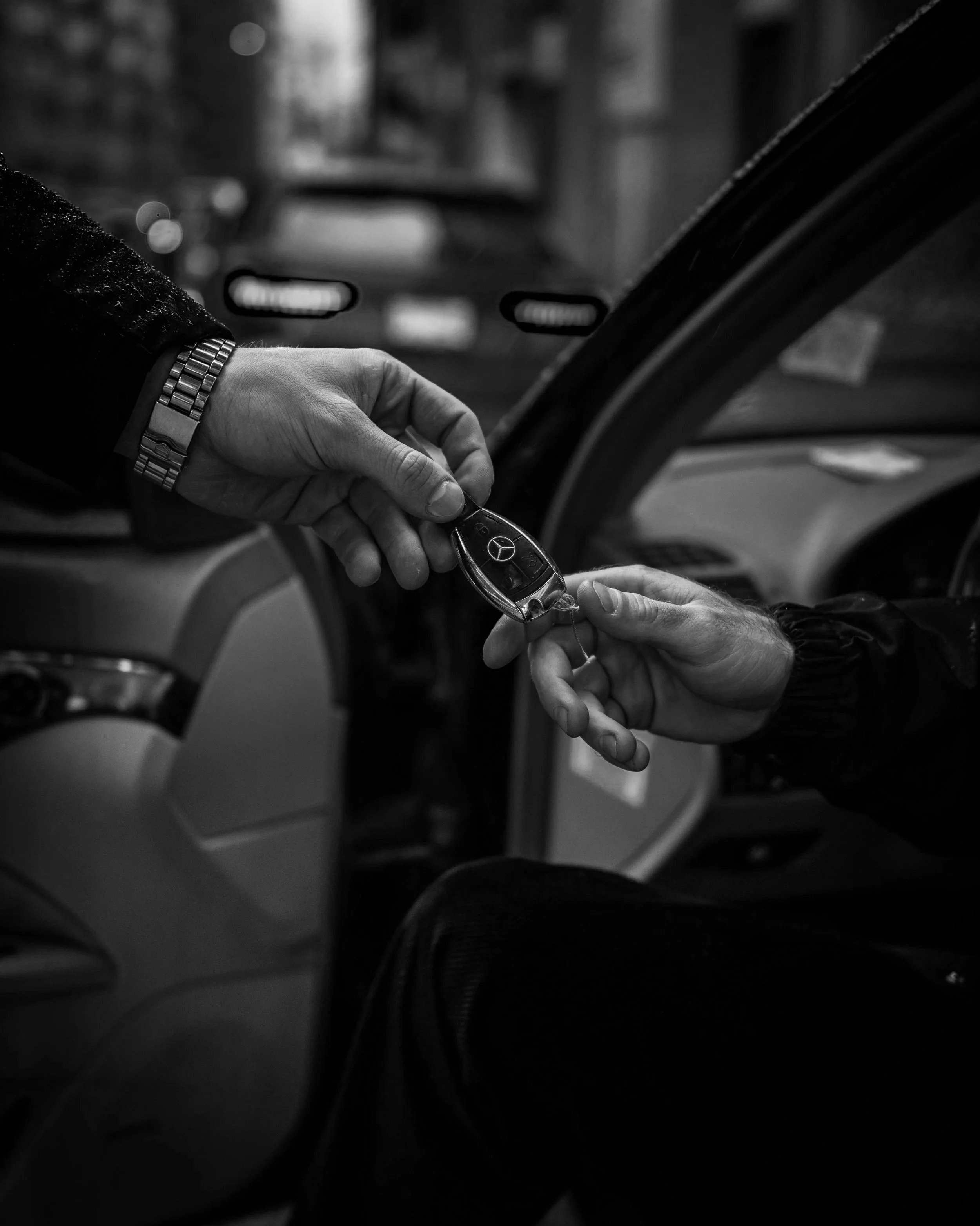 Black and white photo of two hands exchanging a Mercedes-Benz car key inside a vehicle.