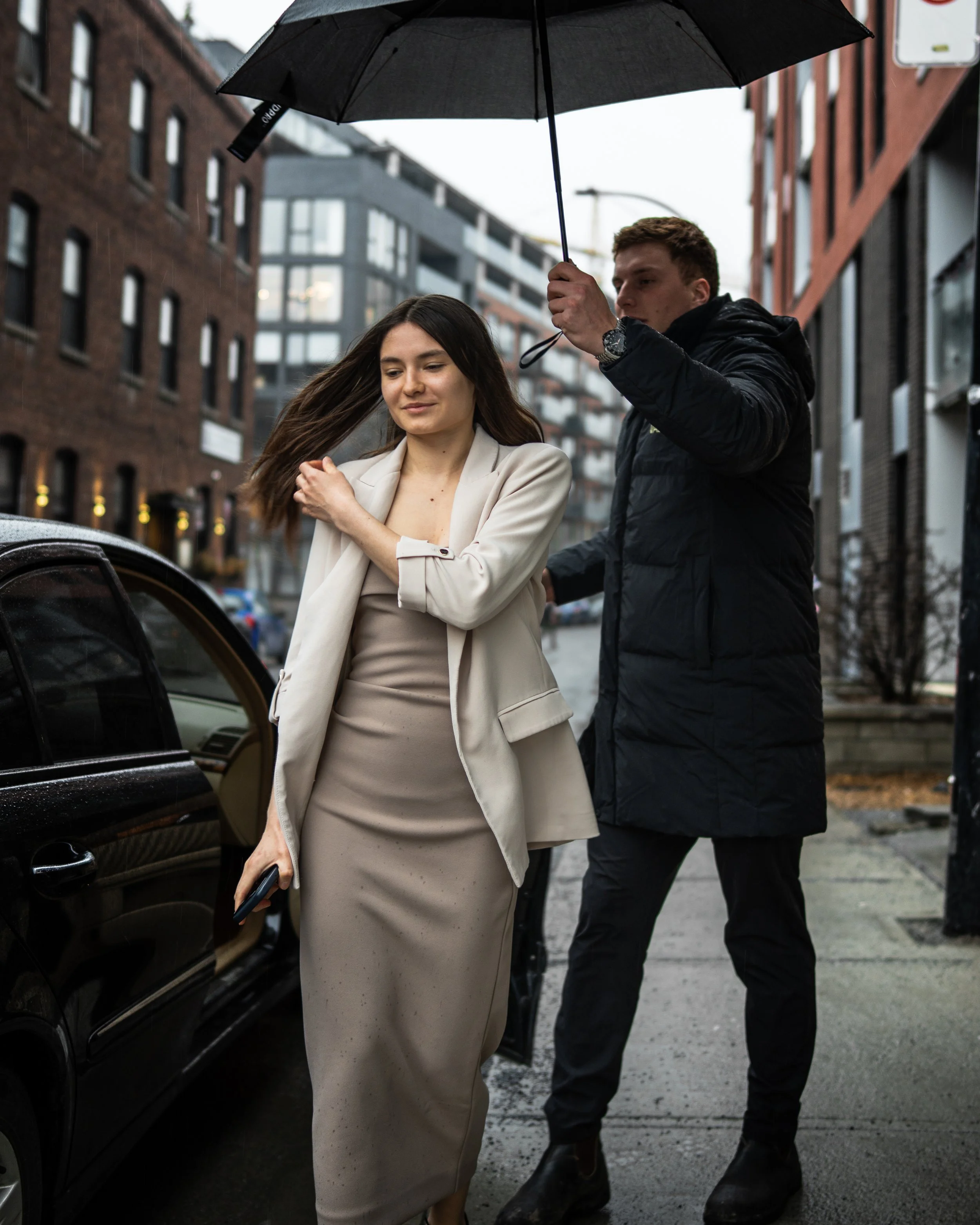 A woman with long brown hair wearing a beige blazer and skirt, holding a small black object, is being assisted by a man with short brown hair in a black jacket, who is holding a black umbrella over her on a rainy city street.