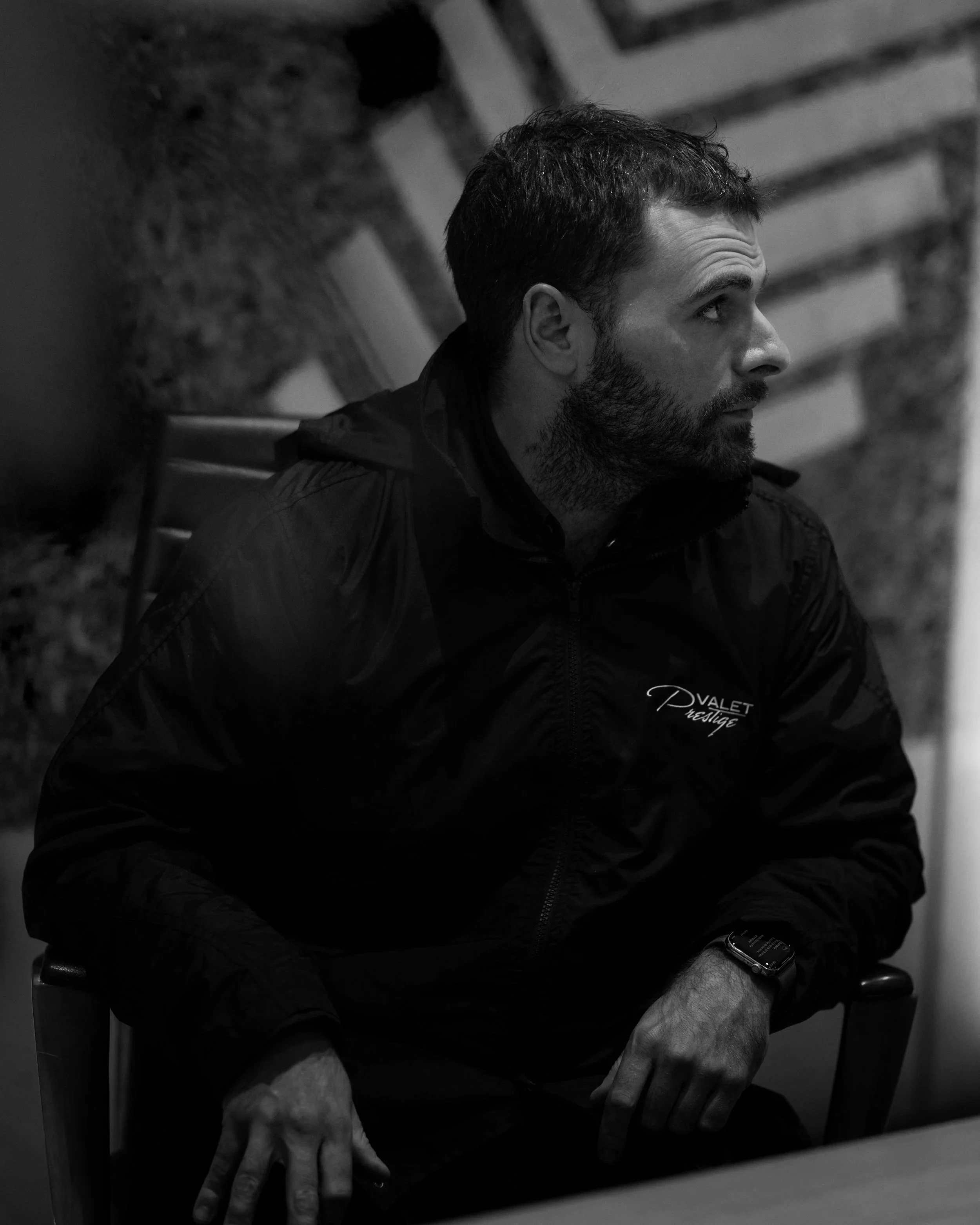A man with dark hair, beard, and mustache sits on a chair, looking to his right. He is wearing a black jacket with a logo on the chest and a watch on his left wrist. The background shows blurred objects, possibly framed pictures or artwork.