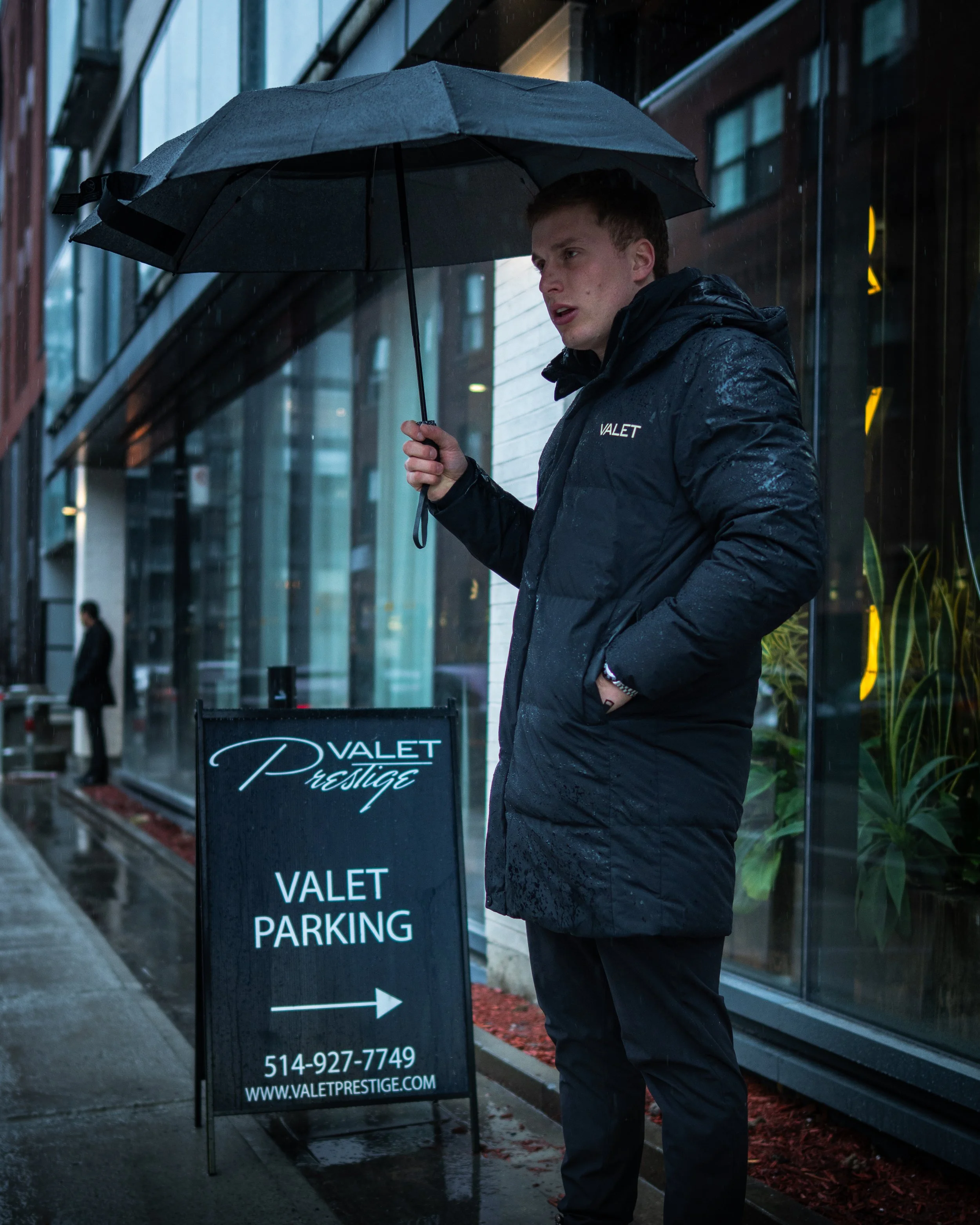 A young man in a dark rain jacket standing outside in the rain under an umbrella beside a valet parking sign. The parking sign displays the name 'Valet Prestige,' phone number, and website. Backdrop includes a glass building facade and a sidewalk with wet pavement and red mulch.
