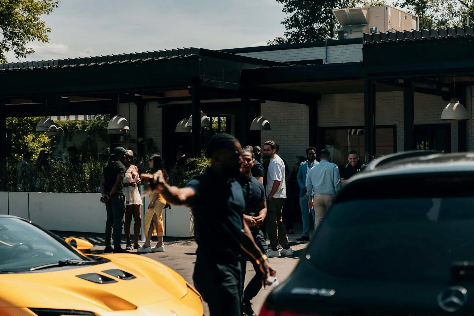 People gather outside a building with luxury cars parked in front, some chatting and socializing.