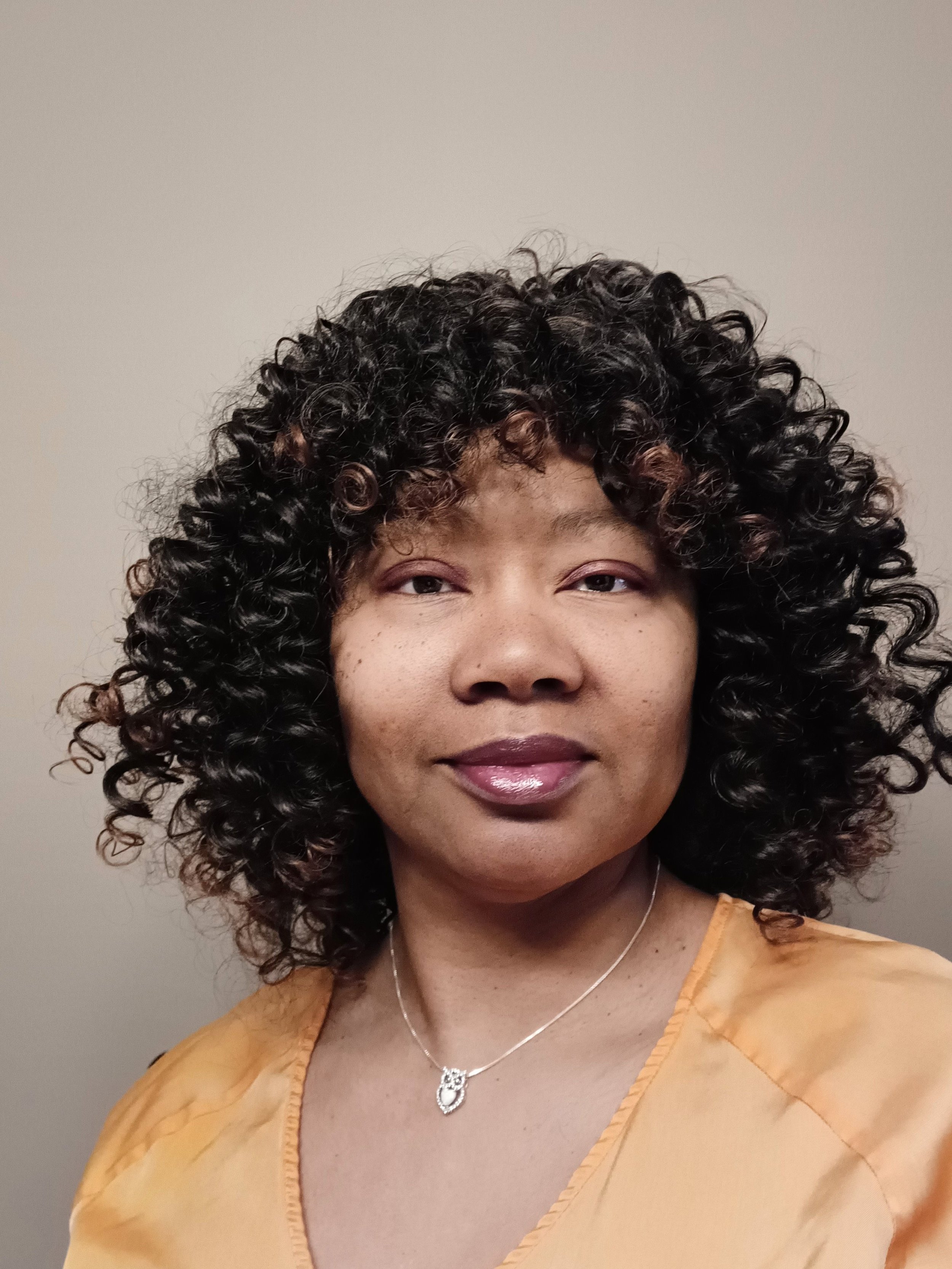 A woman with dark, curly hair wearing a yellow top and a silver necklace with a pendant.