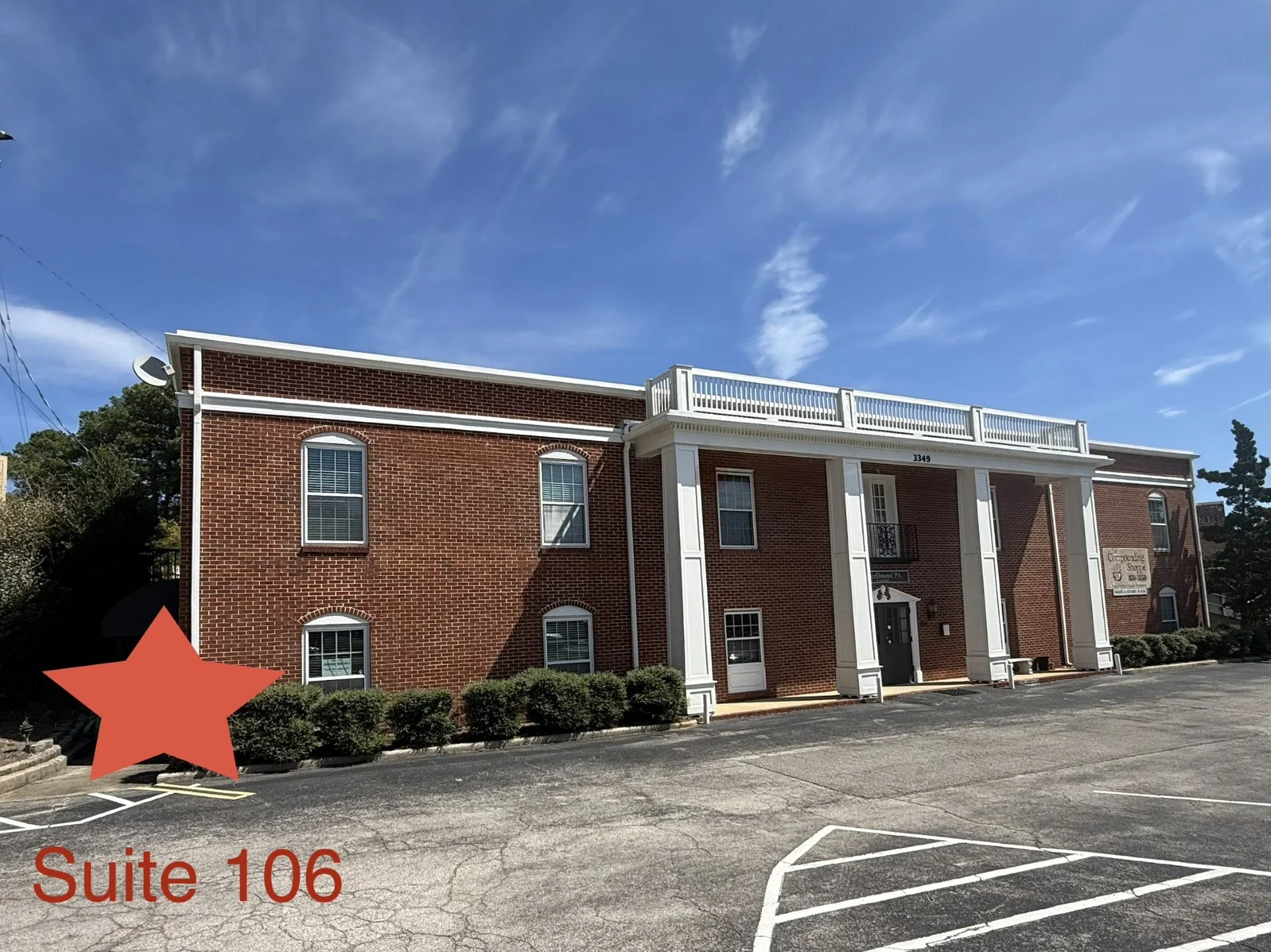 Red brick building with white columns, windows, and railing balcony under a blue sky with wispy clouds. Parking lot in foreground with marked parking spaces and a red star graphic. Text alert 'Suite 106' at the bottom left corner.