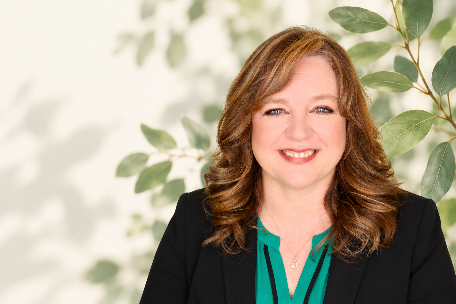 A woman with shoulder-length curly red hair, blue eyes, and a friendly smile, wearing a black blazer and a green blouse, standing in front of a leafy background.