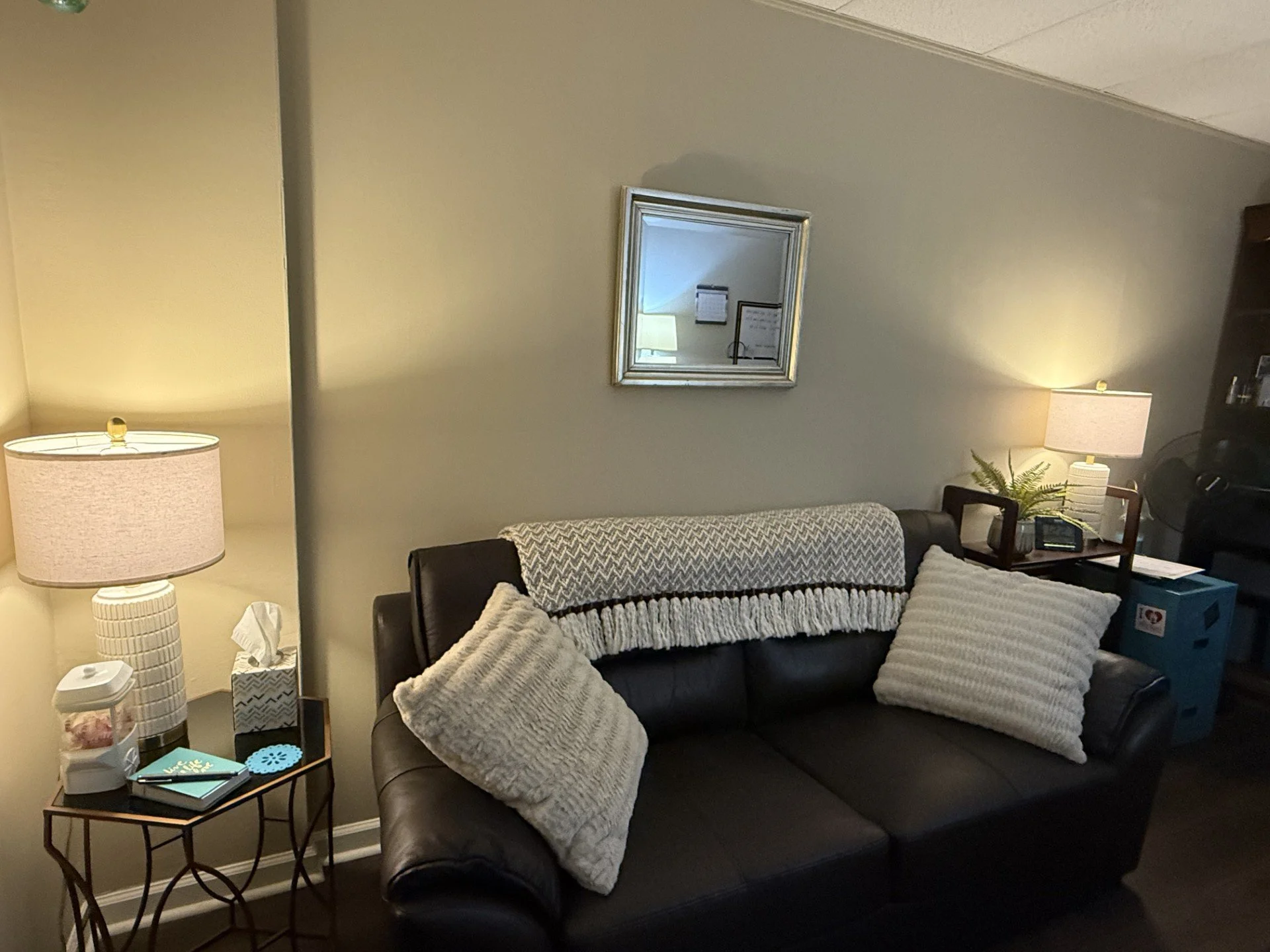 Living room with a dark brown leather sofa decorated with a white knitted blanket and striped pillows, a small round side table with books, a lamp, a tissue box, and a framed mirror on a light-colored wall, with a dark wooden bookshelf on the right side.