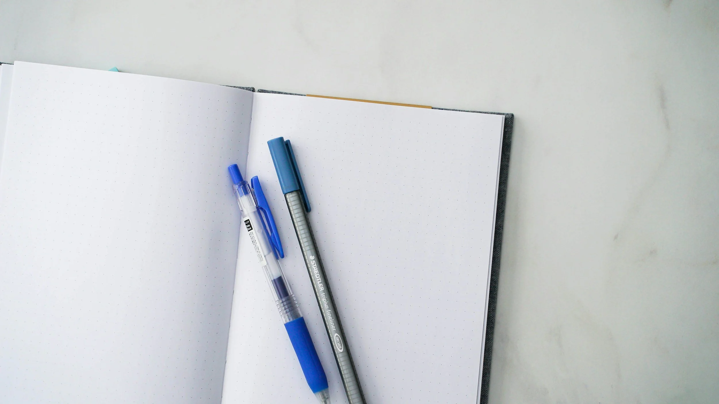 Open dotted notebook with three pens resting on it, on a white surface.