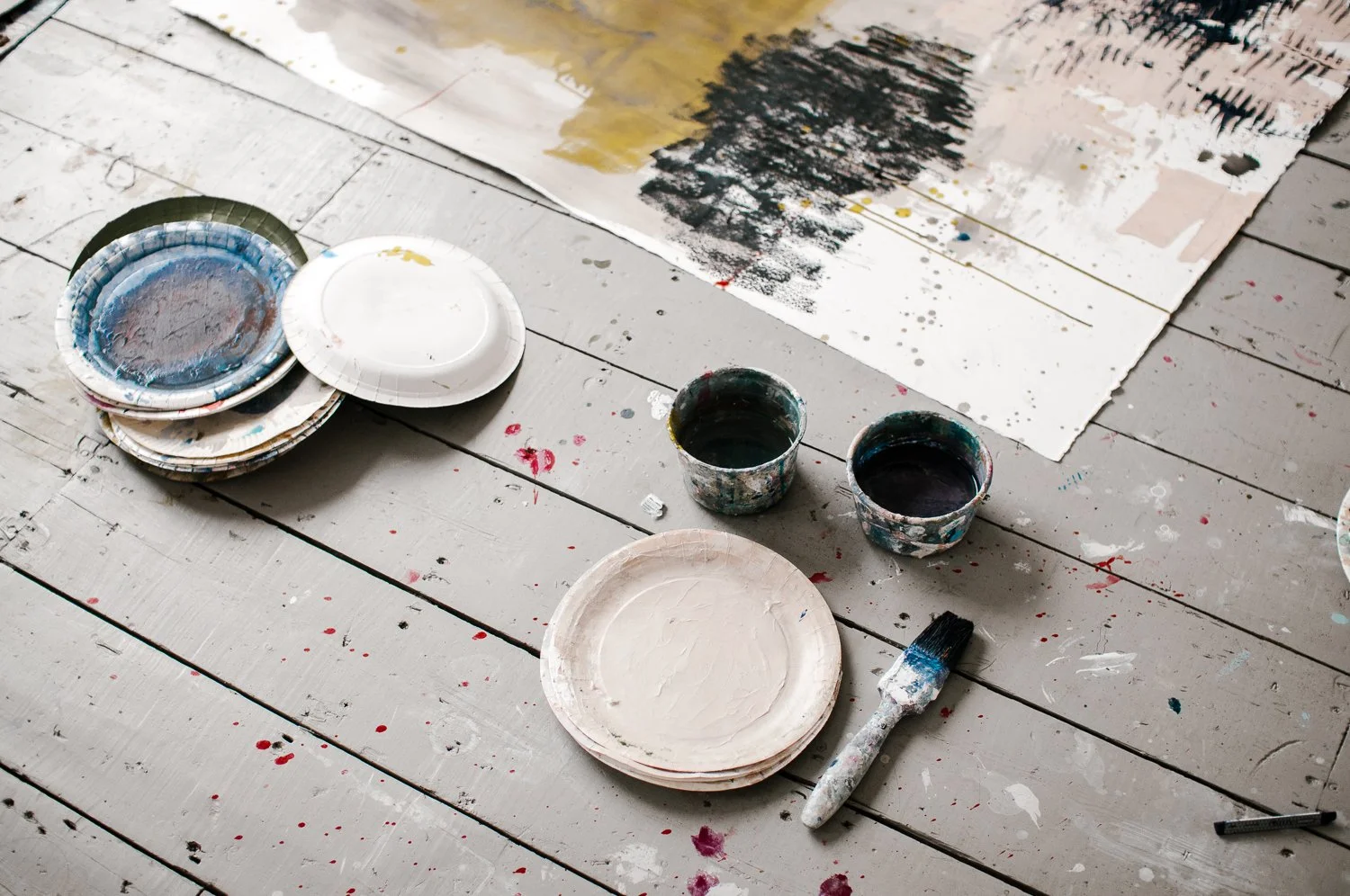 Paint supplies including stacked paper plates with paint, cups with dark paint, a paintbrush with white and blue paint, and a work of abstract artwork on a white wooden floor that has splatters of paint.