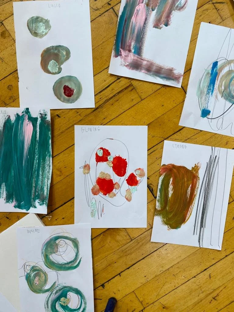 Collection of children's abstract paintings on white paper scattered on a wooden floor, using various colors and brushstrokes.