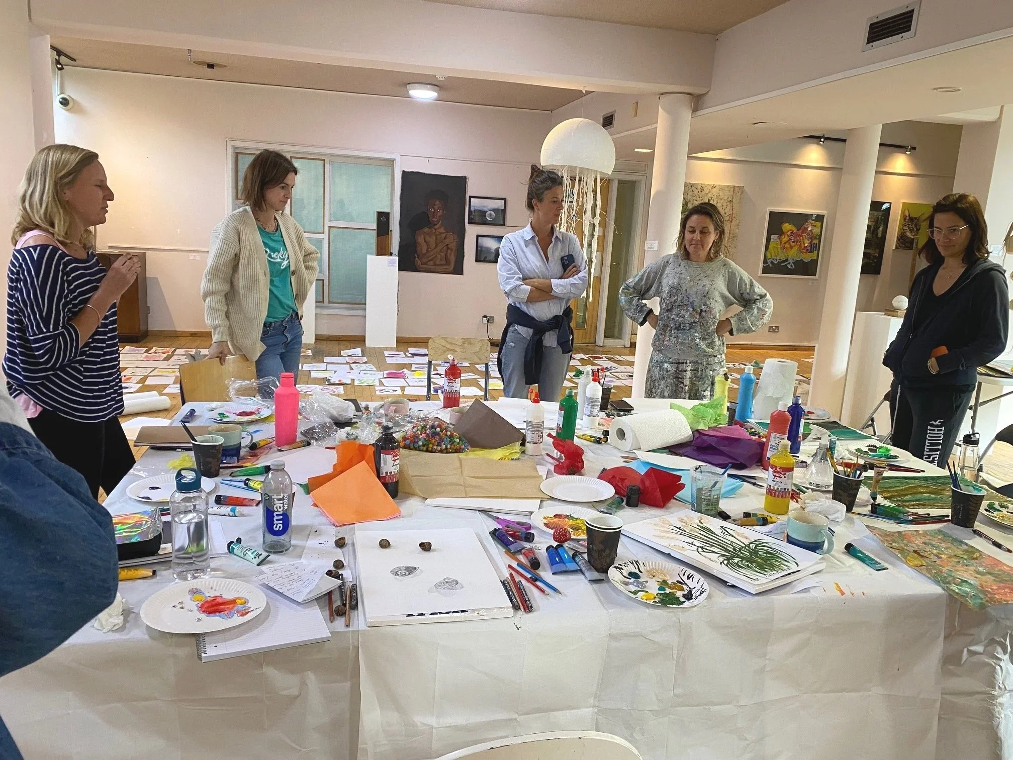 An art gallery or studio space with five women standing around a table filled with art supplies and projects, some artwork on the walls, and a large jellyfish-shaped light fixture hanging from the ceiling.