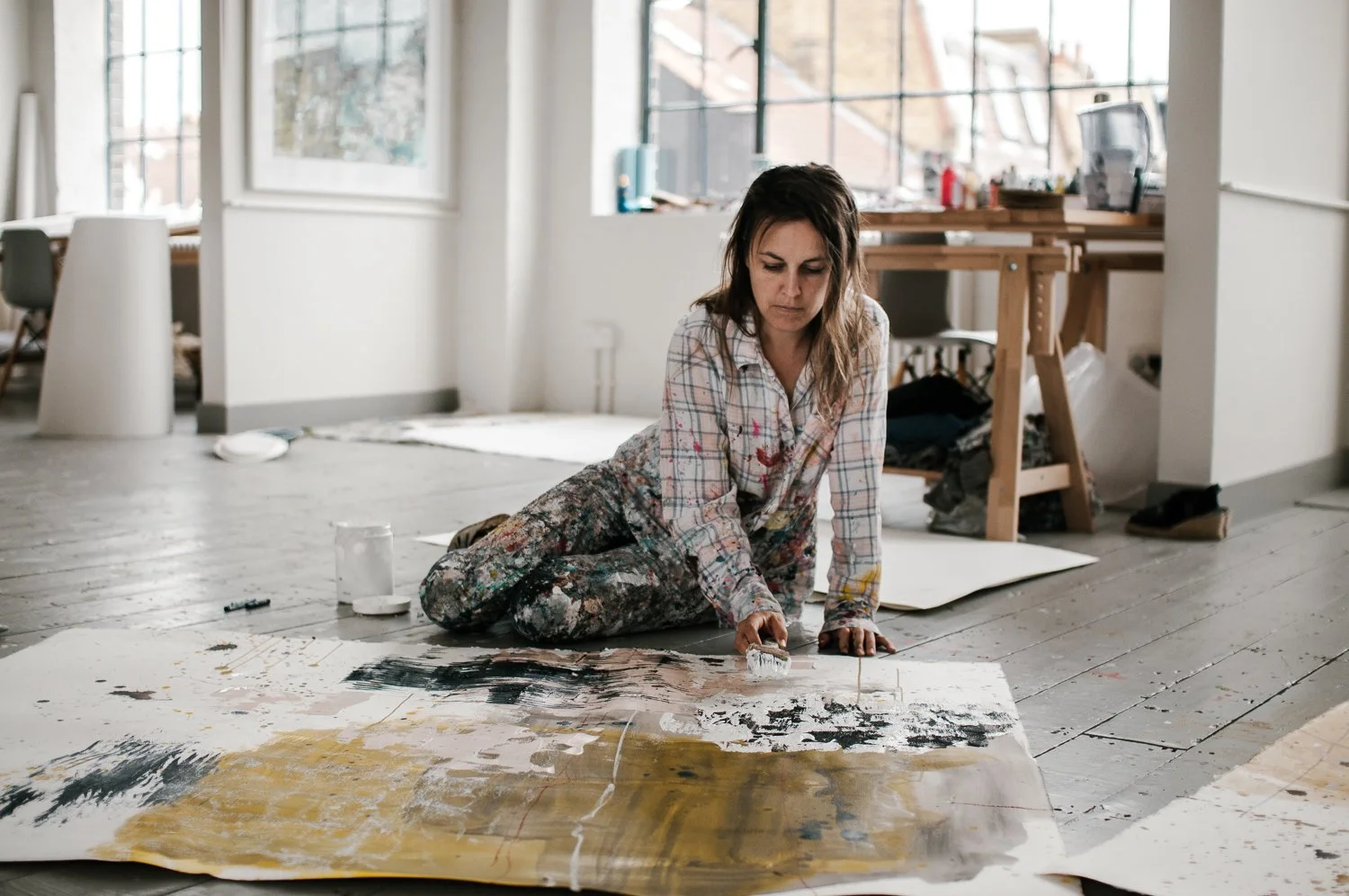 Artist Harriet Hoult in paint-splattered clothes is kneeling on the floor, using a roller brush to paint on a large canvas laid on the wooden floor of a bright, spacious studio. The background shows large windows with a view of buildings.