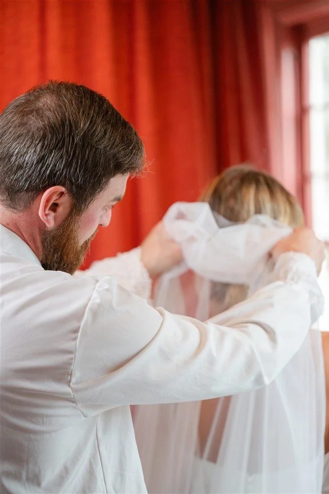 The Bridal Steward helping a woman put on a white veil in a room with red curtains.