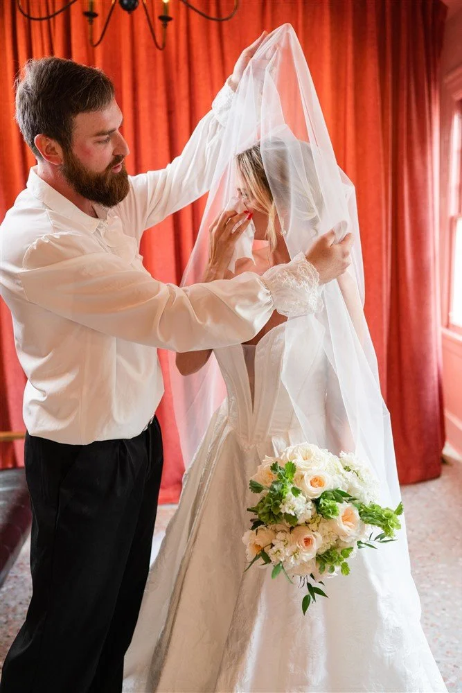 The Bridal Steward adjusts a bride's veil inside a room with red curtains and a window, with a bouquet of white and blush pink roses in her hand.