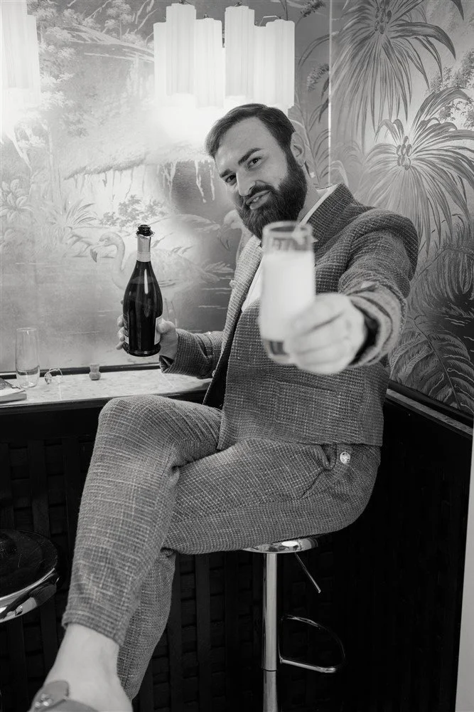 The Bridal Steward in a suit sitting on a barstool, holding a bottle of champagne in one hand and a champagne glass in the other, smiling and pointing the glass towards the camera, with a tropical mural in the background.