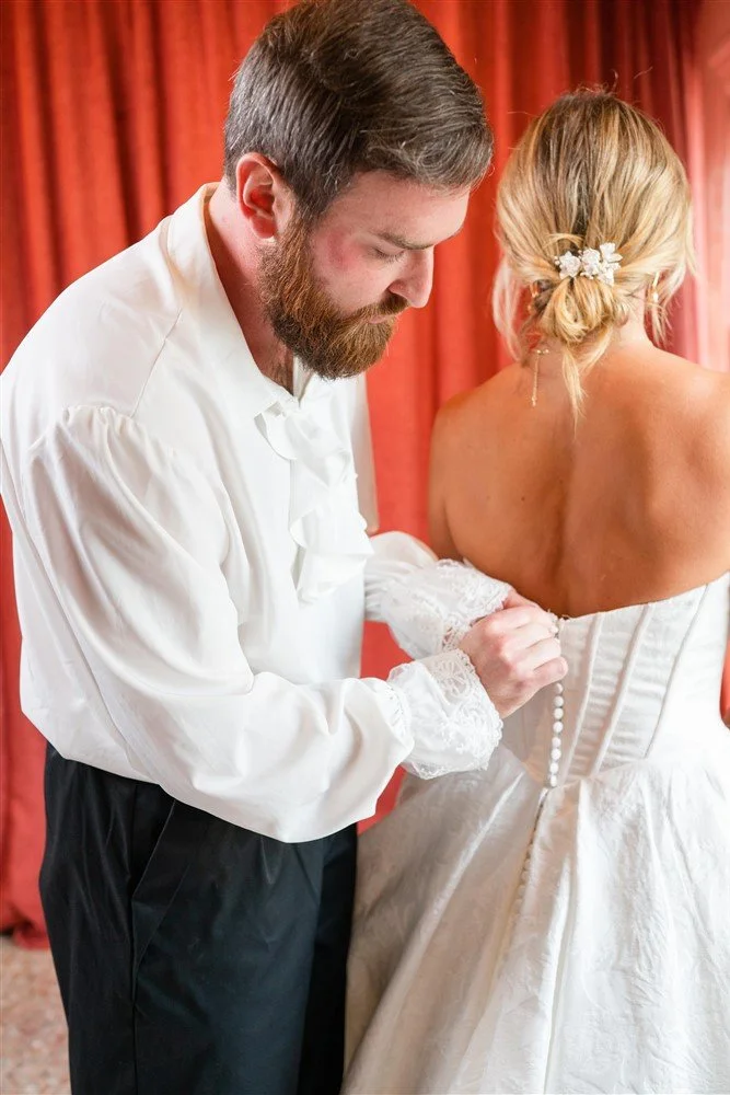 The Bridal Steward helping a woman into a wedding dress, with red curtains in the background.